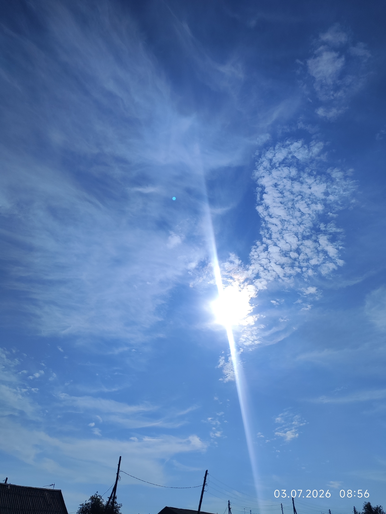 This is a vertical, outdoor, daytime photograph taken in Konovalovo, Russia. The image is dominated by a bright blue sky filled with wispy white clouds and a prominent sunbeam diagonally crossing the frame from the bottom left towards the top right. The sun itself is highly illuminated and partially obscured by clouds, creating a lens flare effect. In the lower portion of the image, a dark roofline of a building is visible on the left. Electrical poles and wires are also present, stretching across the bottom edge of the frame. A date and time stamp in white text reads "03.07.2026 08:56" in the bottom right corner.