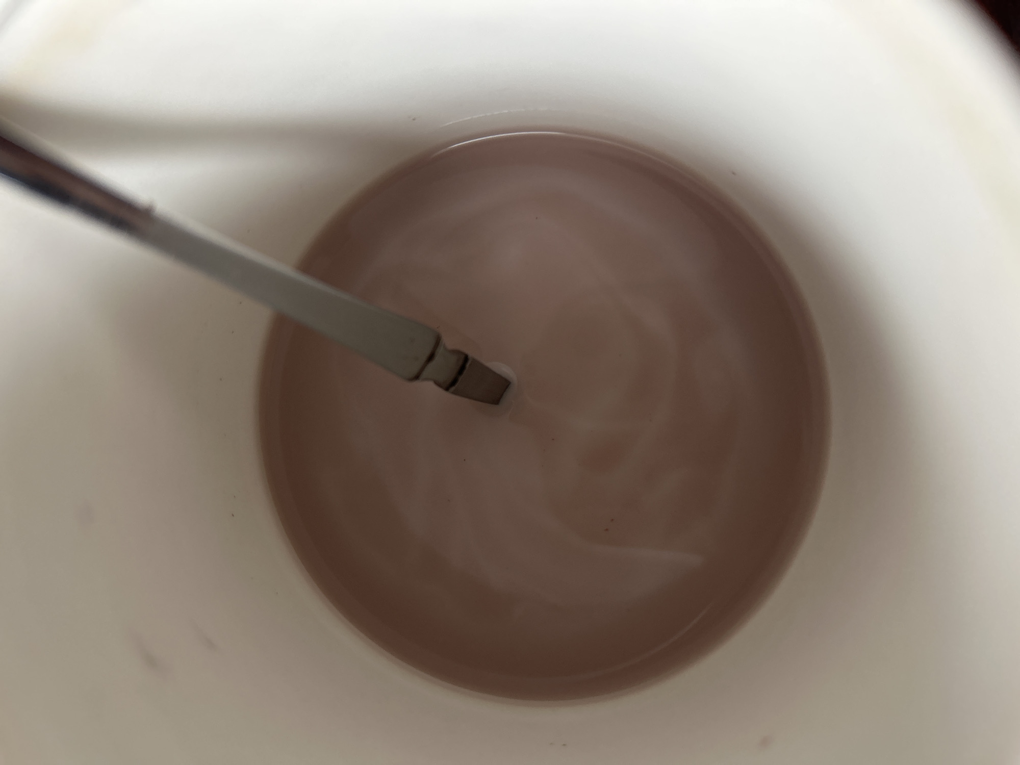 A top-down, close-up shot shows a white mug filled with a light brown liquid, possibly hot chocolate or coffee with milk. A silver spoon is partially submerged in the liquid, with a visible swirl pattern on the surface of the drink, suggesting it has been recently stirred. The mug appears to be on a flat surface, and the lighting is soft and diffused. The background outside the mug is out of focus and appears to be a lighter color, possibly a table or countertop. The image does not provide any specific details about the city of Kaa-Khem, Russia, or any individuals present.