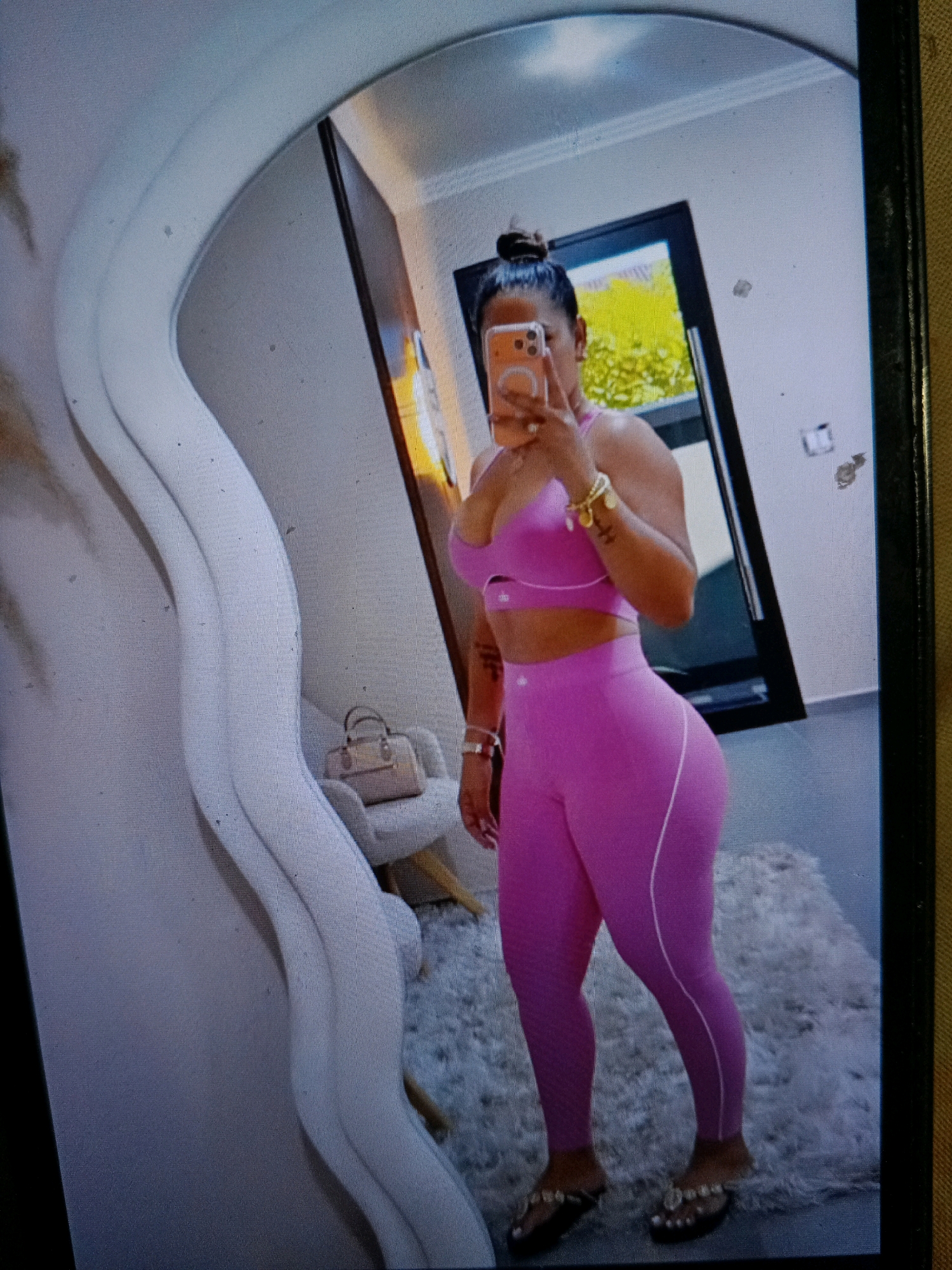 A full-length mirror reflects a person wearing a pink athletic set, consisting of a sports bra and leggings. The person is holding an orange smartphone in their right hand, as if taking a selfie. The background shows a room with a doorway, a window, and a fluffy rug. The scene is located in a residential setting in Noord, Suriname.