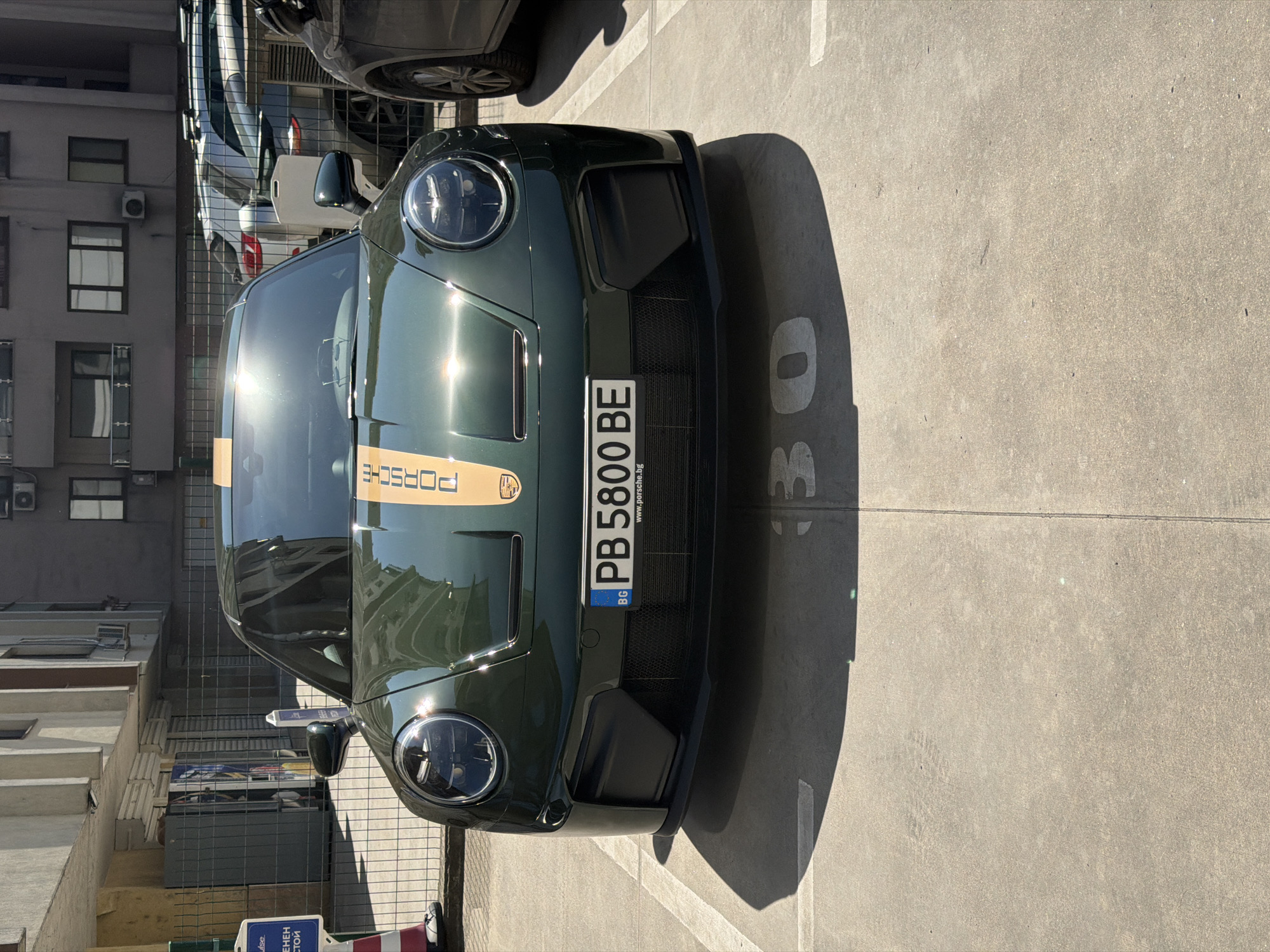 The image shows a dark green Porsche 911 parked in a parking space marked with the number "30". The license plate reads "PB 5800 BE" and indicates registration in Bulgaria ("BG"). The car has a gold stripe down the hood with the Porsche logo. The surrounding environment appears to be a city parking lot with a building visible in the background, with windows, and possible air conditioning units. The location is most likely Plovdiv, Bulgaria based on the license plate.