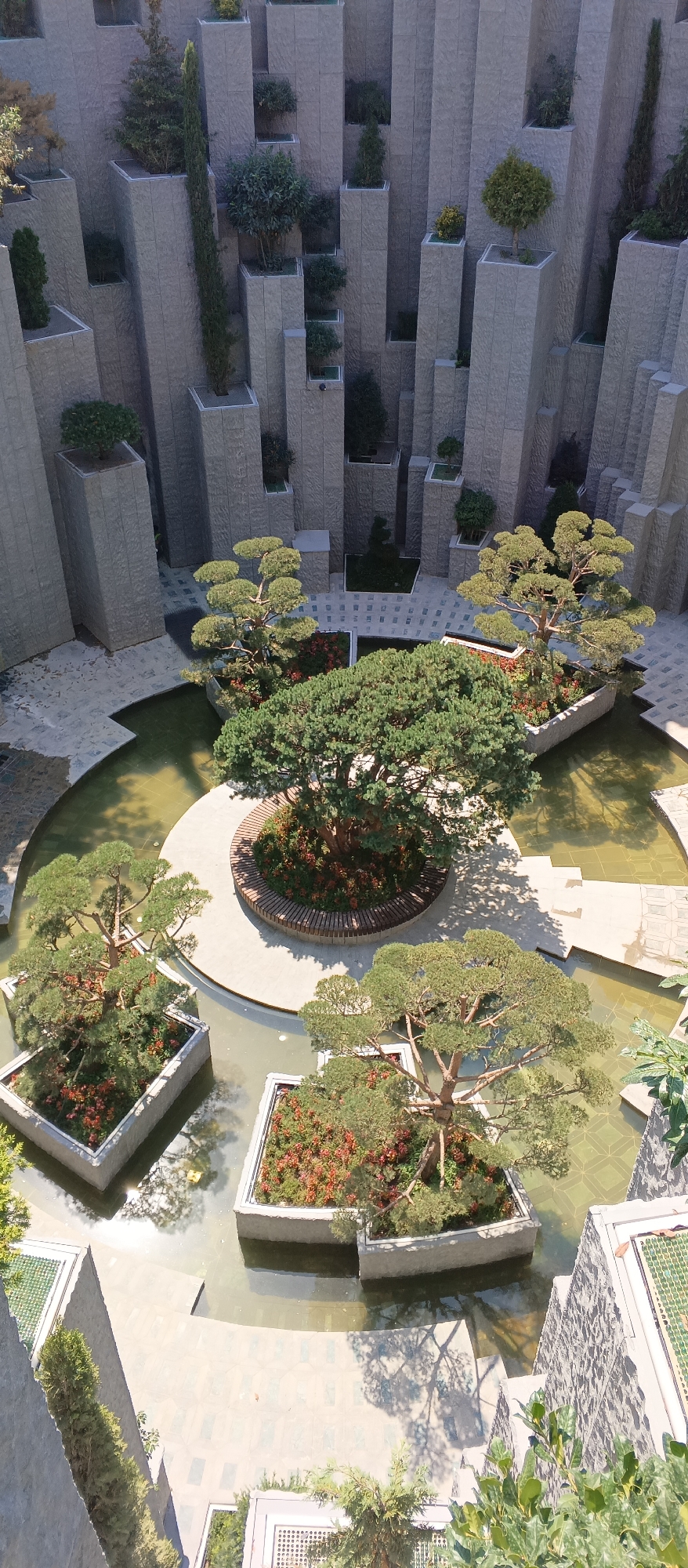 This is a high-angle, full-shot, eye-level, outdoor, daytime, daylight, clear-weather photograph of a stylized garden. The garden features several large, rectangular planters, some partially submerged in water, each containing a bonsai tree. One large bonsai tree is centrally located in the image, surrounded by a circular wooden bench. The planters and surrounding pathways are made of light-colored stone, with some areas featuring patterned tiling. Tall, slender, grey stone pillars of varying heights are arranged around the perimeter of the garden. The pillars have a rough, textured surface and are topped with small plantings. The overall aesthetic is modern and minimalist, with an emphasis on geometric shapes and natural elements. There are no people visible in the image. The lighting suggests a sunny day, casting distinct shadows from the trees onto the ground and water.