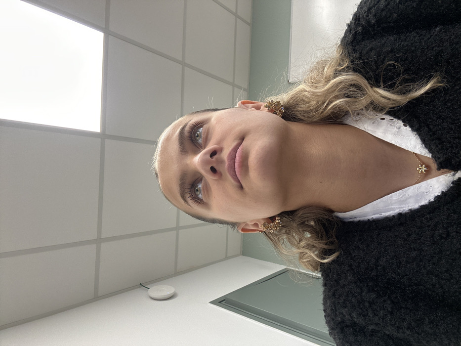 The image displays a head-and-shoulders portrait of a female subject, observed from a low-angle perspective, situated indoors in what appears to be a modern institutional or office environment in Saint-Étienne, France. The subject, a young woman, looks upward with a neutral expression. She has light-colored eyes, prominent eyelashes, and light brown or blonde curly hair visible around her ears and shoulders. She is wearing a black textured cardigan over a white collared shirt with eyelet details. Gold-toned floral or clustered earrings are present on both ears, and a gold-toned necklace with a small, flower-shaped pendant rests at her collarbone. The background consists of a white wall on the left, where a white circular device, possibly a sensor or detector, is mounted near a thin dark cable. A light green door frame or panel is partially visible on the far left. The ceiling is a white suspended grid system with several rectangular, illuminated LED light panels casting bright white light.