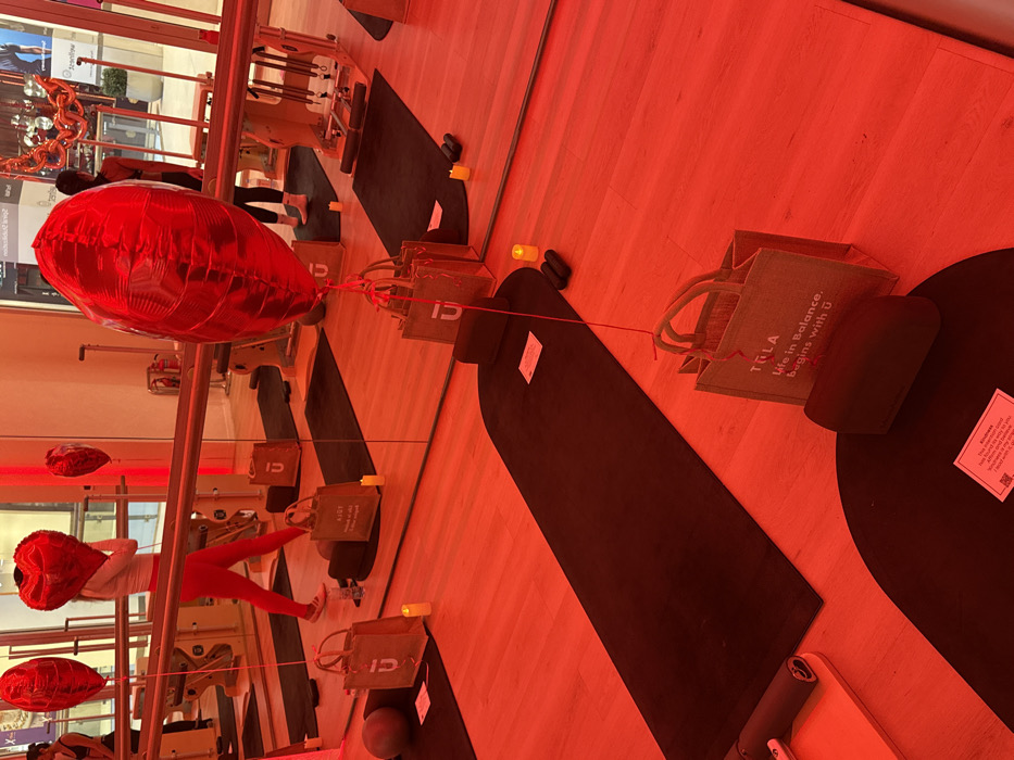 The image portrays an indoor fitness studio, likely a barre or Pilates facility, located within a commercial complex in Dubai, United Arab Emirates. The room is uniformly illuminated with intense red artificial lighting, casting a reddish hue across the light wood floor and mirrored walls. Multiple black yoga mats are systematically arranged on the floor.
Each mat station includes a grey-brown canvas tote bag labeled "TULA Life in Balance. begins with U," a red heart-shaped foil balloon tied to the bag with a red string, and one or more small, battery-operated LED candles emitting a warm, yellowish glow. Additional fitness equipment such as black foam rollers and small black dumbbells are present near some mats. A grey exercise ball rests near a mat on the far left. Small white cards, one visibly reading "Kindness" with further text, are placed on some mats. A ballet barre extends horizontally along the mirrored wall.
Two individuals are visible, primarily in reflection. One person, wearing red leggings and a light-colored top, is walking from left to right, holding a red heart-shaped balloon in front of their face. Another person is partially visible in the reflection on the far right, standing near a Pilates reformer machine. The scene is meticulously prepared, suggesting a class or event setup. The mirrored walls reflect the studio's interior and a glimpse of the exterior of the commercial complex.