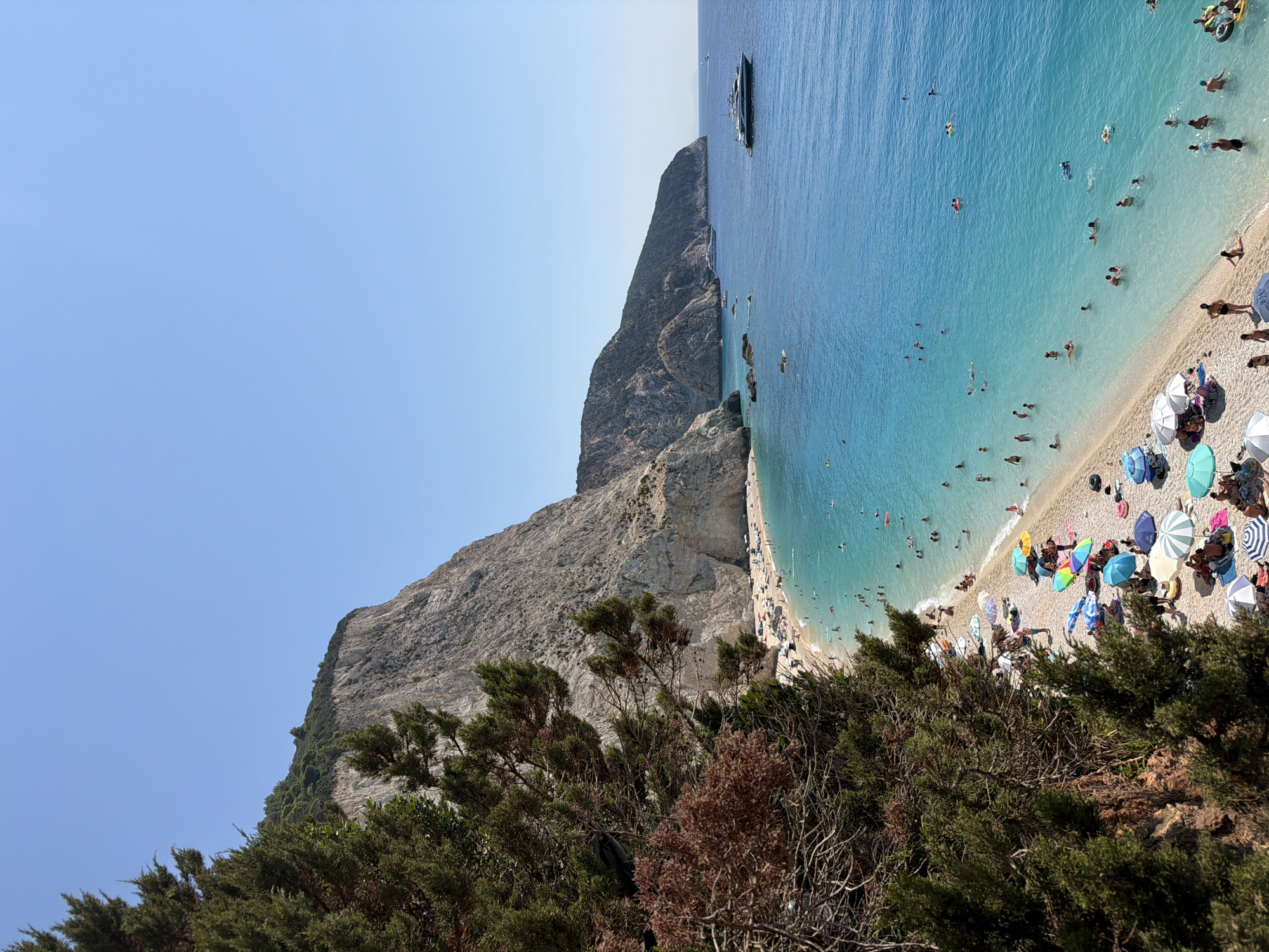 A high-angle, long shot captures a crowded, picturesque beach scene on a bright, sunny day. The turquoise waters of the Ionian Sea gently lap against a pebbled shore, populated by numerous beachgoers. People are scattered across the beach, some lounging under colorful umbrellas, others wading or swimming in the clear water. A large, white yacht is anchored offshore, and several smaller boats are visible in the distance. Towering limestone cliffs, covered in lush green vegetation at their peaks, form a dramatic backdrop to the beach, with a dense cluster of evergreen trees in the foreground on the left side of the image, obscuring a portion of the cliff face and the sky. The scene is located in the Municipal Unit of Apollonioi, Greece.