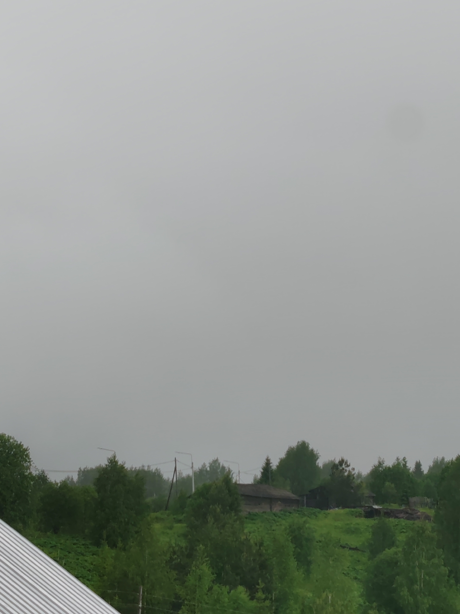 The image presents a landscape on a cloudy day in Чухлэм, Russia. In the foreground, the corner of a corrugated metal roof is visible. Beyond, a gentle slope ascends, covered in lush green vegetation interspersed with trees. Utility poles with wires run across the hillside. Atop the hill, a few structures are visible amidst the greenery, possibly houses or sheds, giving the impression of a rural or semi-rural setting. The sky is uniformly overcast, casting a diffused light over the scene.