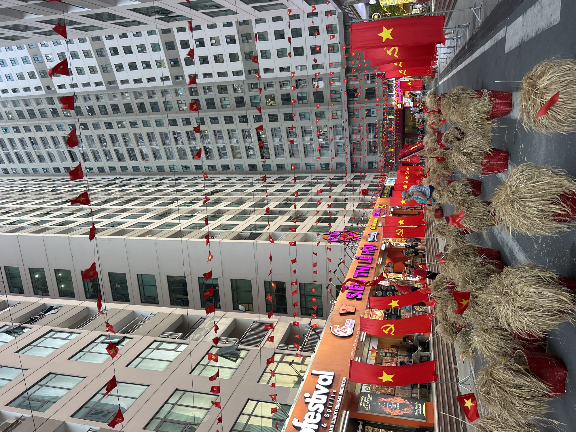 Here is a description of the image:
The image depicts a street in Bắc Nha Trang, Vietnam, adorned with decorations, particularly Vietnamese flags, straw baskets, and banners for a festival. Tall buildings flank the street on both sides, with small Vietnamese flags hanging from lines strung between the buildings. Large red flags with a gold star are displayed along the storefronts, some featuring the communist hammer and sickle symbol. Along the street, decorative straw bundles are placed in red-painted baskets. People can be seen walking and interacting with the storefronts and decorations. The scene suggests a celebration or commemoration is taking place, likely related to a national holiday or event. A storefront called "festival" in Vietnamese (with Russian underneath) is visible, seemingly selling alcohol and spirits.