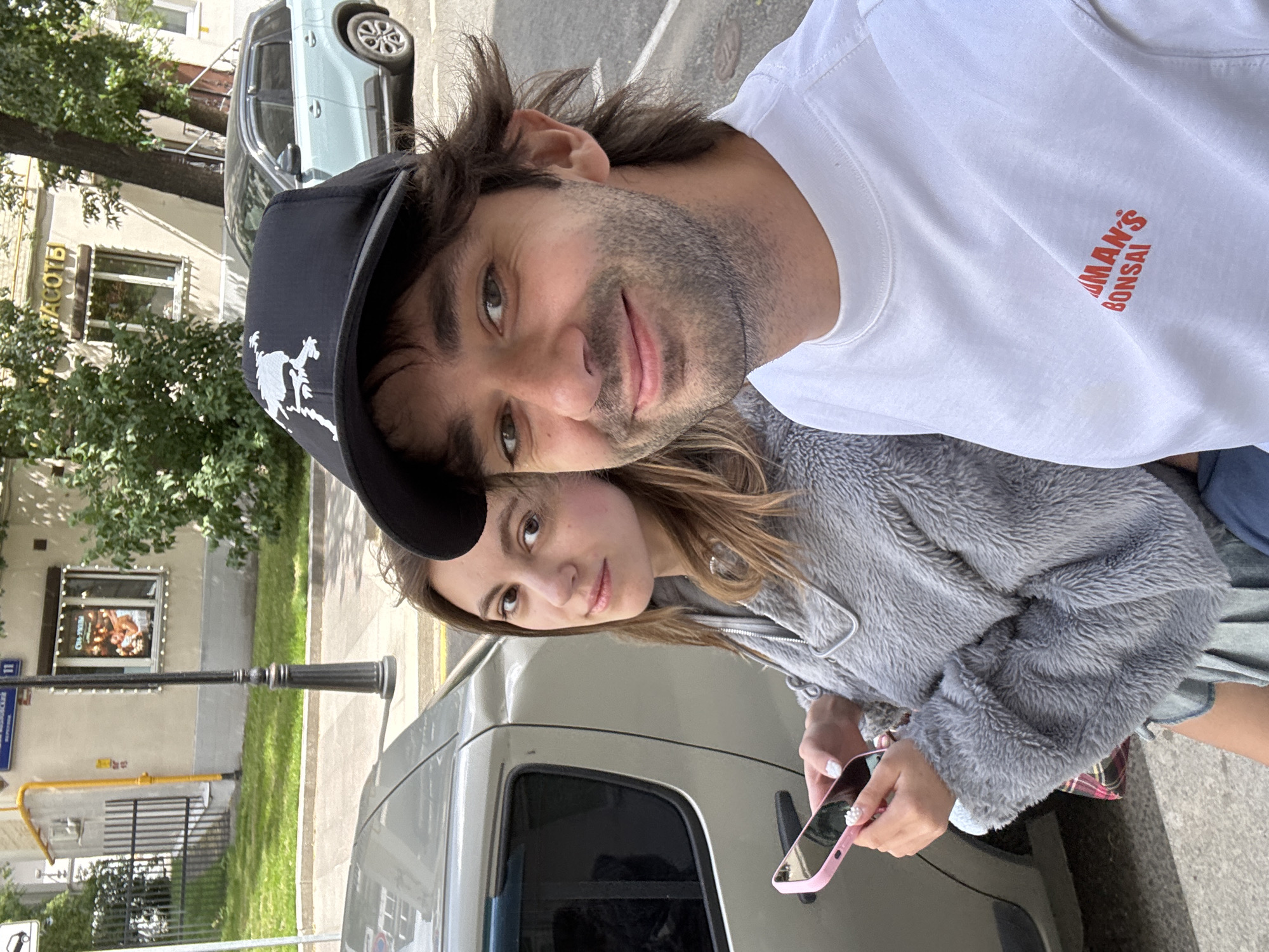 In this selfie, a man and a woman are positioned close together, with the man in the foreground and the woman slightly behind and to his side. The man is wearing a black baseball cap and a white t-shirt with red text that reads "MAN'S BONSAI". He has dark, tousled hair and stubble. The woman has long, light brown hair and is wearing a grey, fuzzy zip-up jacket over a pink and plaid skirt. She is holding a pink smartphone in her right hand.
They appear to be outdoors, with a light-colored car to their right and a building with trees and some greenery in the background. The lighting suggests it is daytime, likely sunny. The building in the background has windows and signs, with one sign displaying text in Cyrillic and an image. A street lamp is also visible. The overall mood seems casual and relaxed, consistent with a selfie taken on a stroll or outing. The location context provided indicates Moscow, Russia.