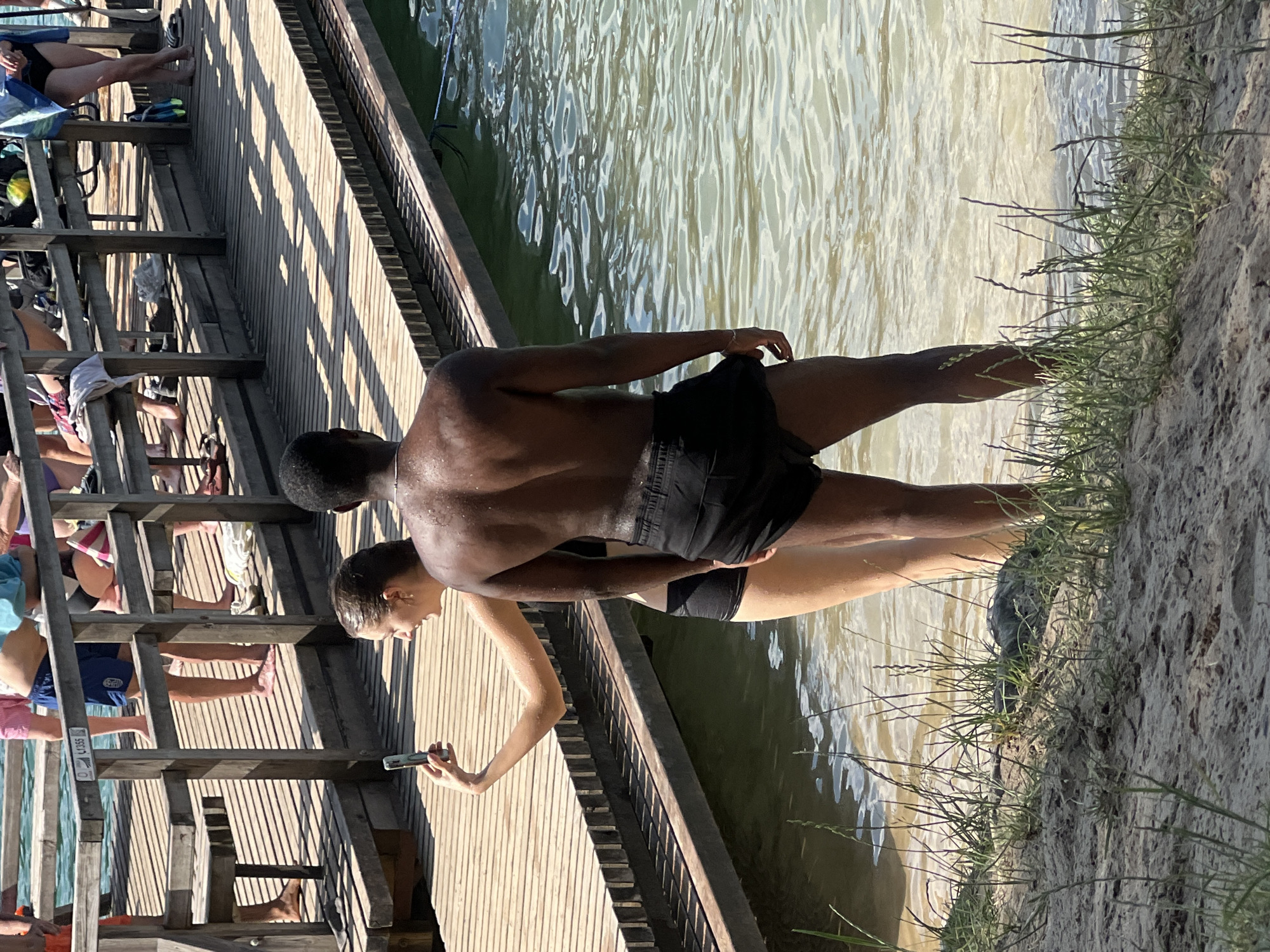 The image shows a group of people on a wooden pier next to a body of water. Two individuals, a man and a woman, are prominently featured in the foreground, standing at the edge of the pier. The man, with dark skin, is partially submerged in the water, with his back to the camera. He is wearing black shorts. The woman next to him, with lighter skin, is also near the water's edge. She is holding a smartphone, seemingly taking a picture. Other people are visible on the pier in the background, some sitting and others standing. The pier is constructed of wooden planks, and some areas have railings. The water appears murky and rippled, suggesting it is a natural body of water like a lake or river. The scene is lit by sunlight, indicating it is daytime. The overall atmosphere seems casual and recreational, likely a summer day at a swimming spot. The setting appears to be a pier in Vilnius, Lithuania. There is a label visible on the pier railing that reads "C1355".