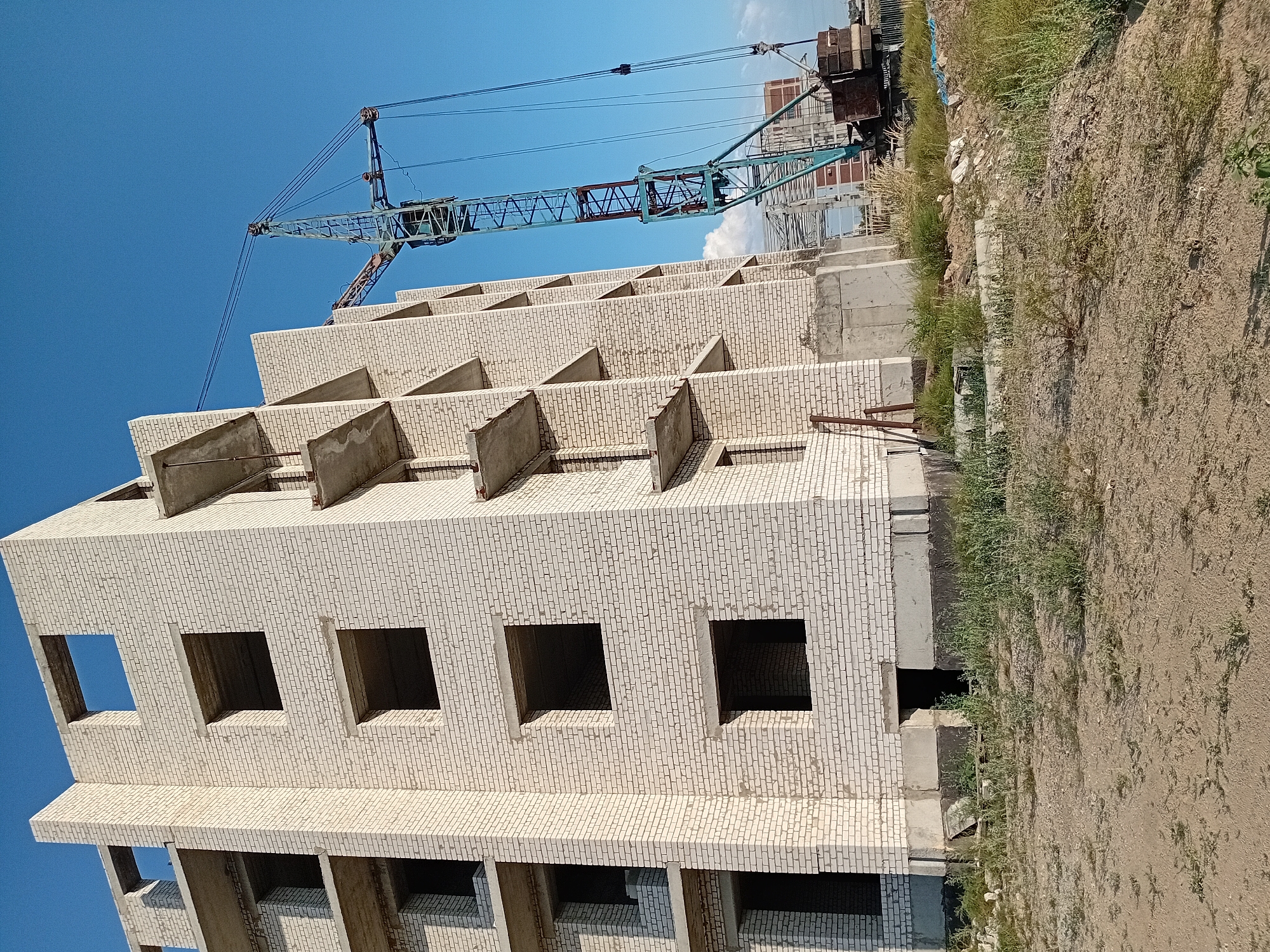 The image shows the unfinished construction of a multi-story brick building in the city of Semey, Kazakhstan. A large blue construction crane with a load is visible to the right of the building. The building has multiple floors with open window and door frames. The surrounding area appears to be a construction site with dirt, scattered debris, and tall grass. The sky is clear and blue.