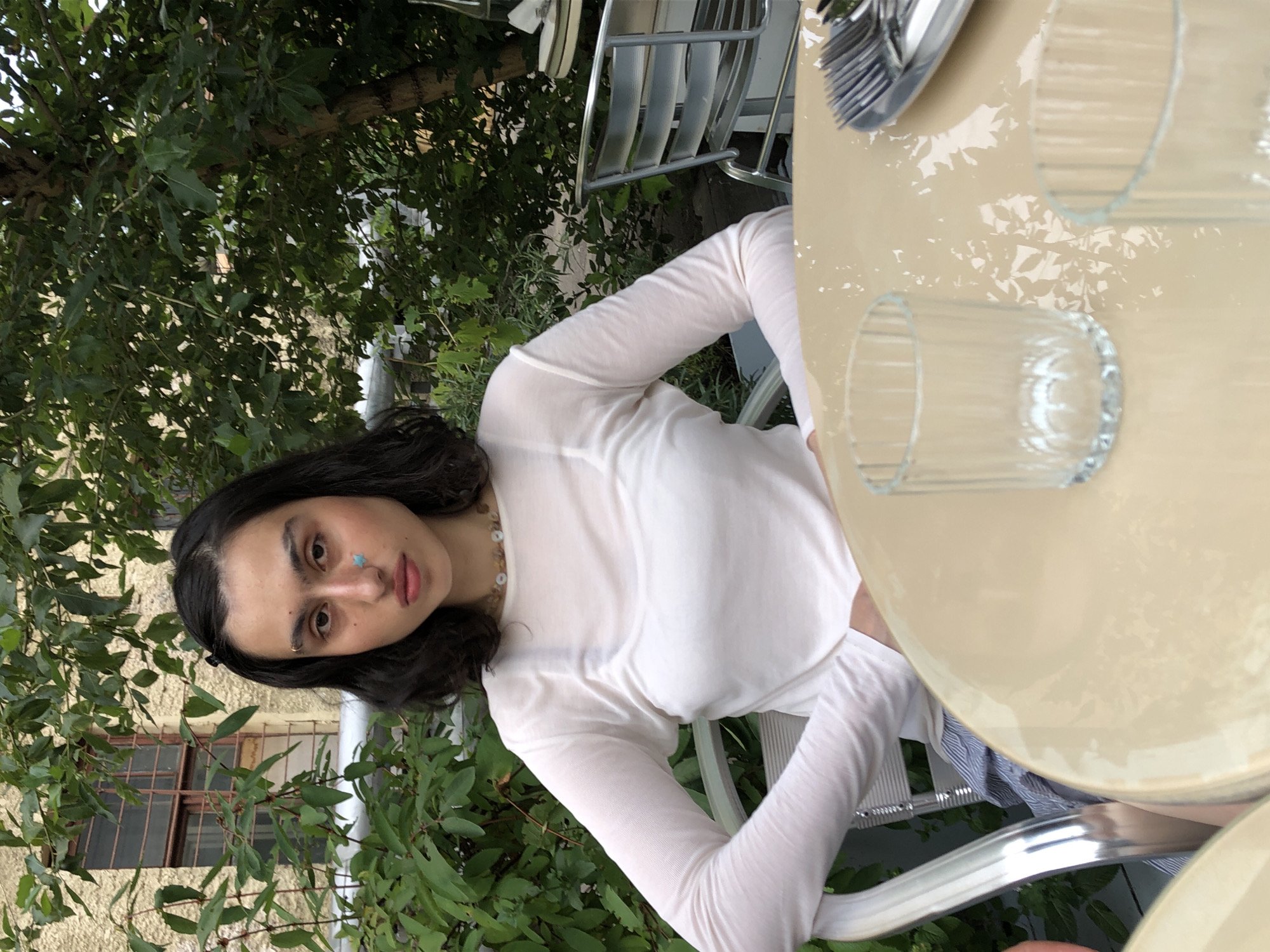 The image shows a woman sitting at a table outdoors. She has dark hair and is wearing a sheer white long-sleeved shirt and a necklace. There is a small blue mark on her nose. She is seated in a chair with a metal frame and a woven back. In front of her is a light-colored, round table with two glasses and cutlery. In the background, there are trees and a building with a window. The scene appears to be in a city, possibly Moscow, Russia.