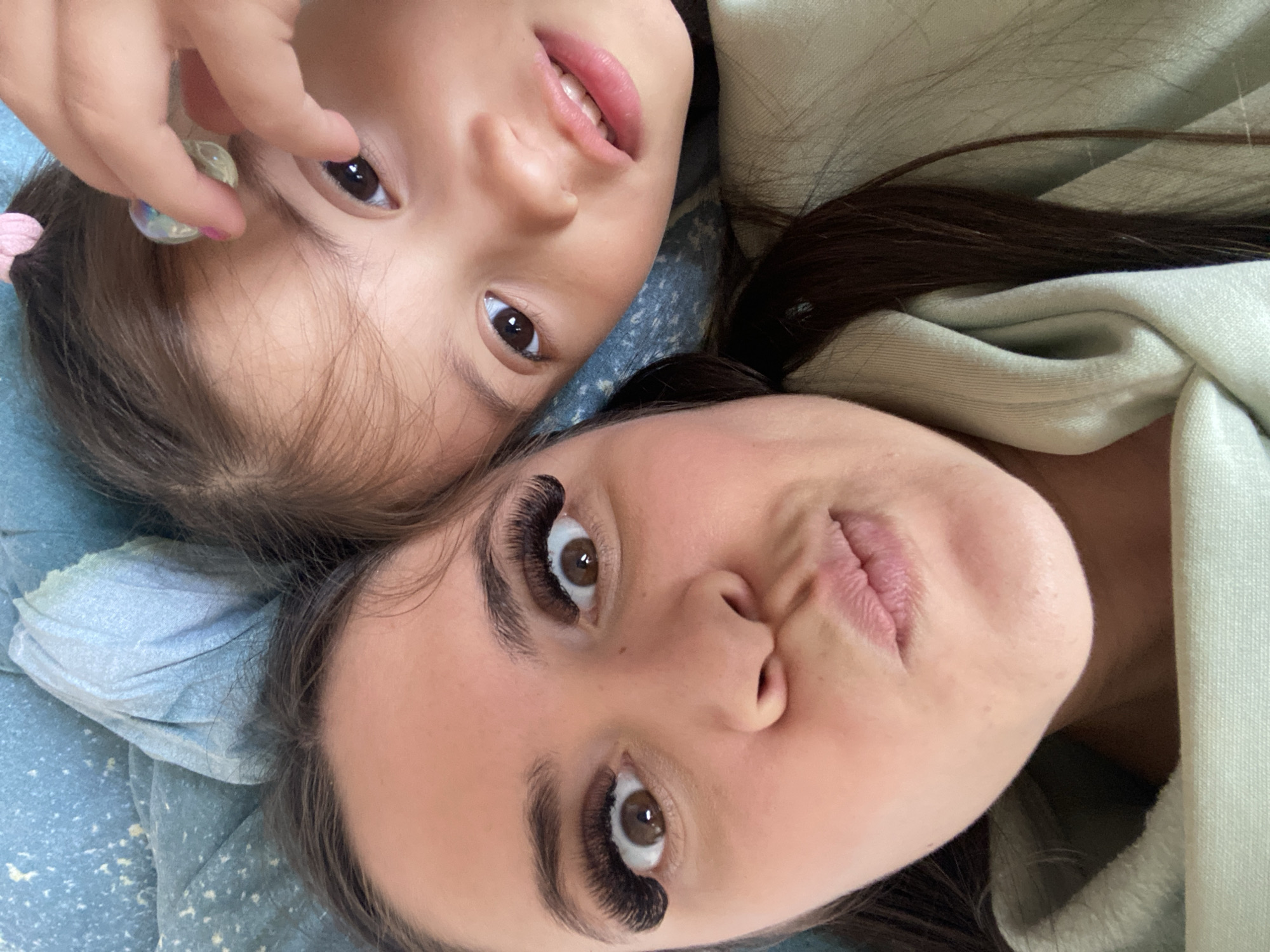 Two individuals, a child and an adult female, are positioned closely together, possibly lying down. The child, located at the top of the frame, has brown hair with a pink hair accessory, brown eyes, and is holding a marble near her face. The adult female, at the bottom, has long, dark brown hair, brown eyes with prominent eyelash extensions, and light skin. They are both looking directly at the camera. The background includes a light blue fabric with a mottled pattern and a light green garment. No evidence suggests this image was taken in Zhigalki, Russia.