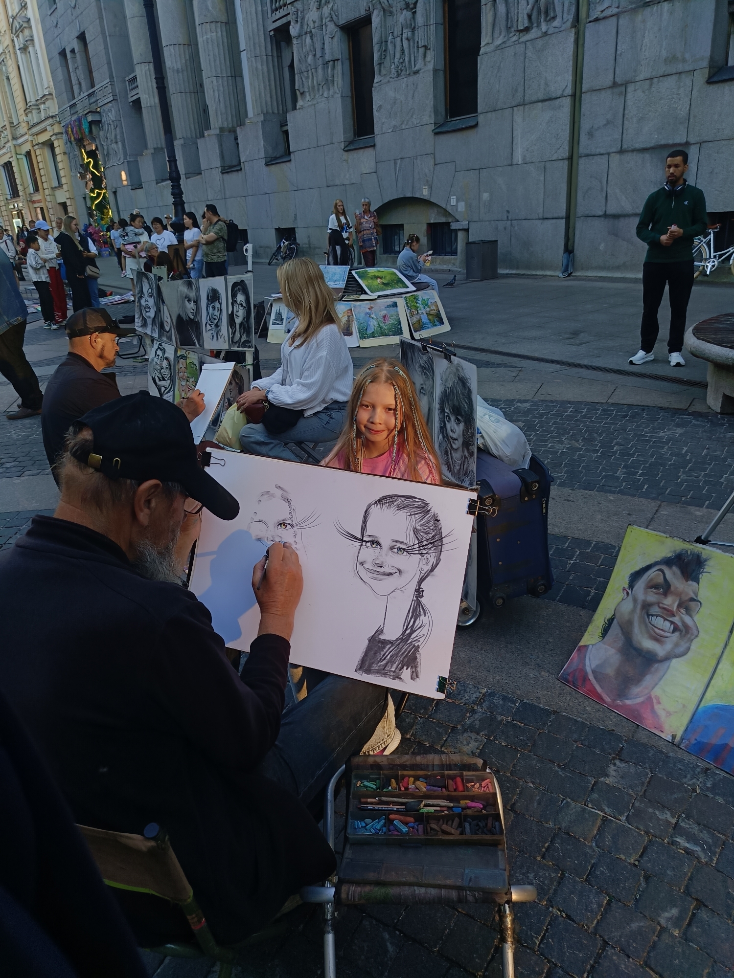In the heart of Saint Petersburg, Russia, an outdoor artist is captured in the act of drawing a caricature of a young blonde girl. The scene unfolds on a cobblestone street, with a grand, classical building serving as a backdrop. The artist, a man with grey hair and a beard, is seated, intently focused on his work, his hand holding a drawing utensil over a large sketchpad. The sketchpad displays a partially completed caricature of the girl, exaggerated and humorous in style.
Beside the artist's sketchpad, the young girl, who is the subject of the caricature, sits with a bright smile, her blonde hair adorned with subtle braids. To her left, a series of finished portraits are displayed, showcasing various faces drawn in a similar artistic style. Further to the right, a vibrant yellow canvas features a completed caricature of a smiling man, his features boldly defined.
In the background, other artists and their displays are visible, as are several onlookers strolling along the street. The overall atmosphere suggests a lively cultural hub, a place where artists gather to showcase their talents and engage with the public in the historic setting of Saint Petersburg.