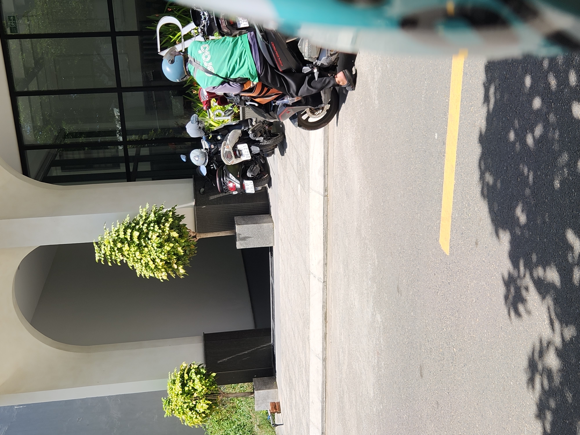 The image depicts a street scene in Da Nang, Vietnam, in front of a modern building with a large arched entrance and glass facade. Two parked motorcycles are visible. One is a black scooter with a license plate "79-01 934.24". Another black scooter, partially obscured, has a license plate "06221". In the foreground, a person wearing a bright green Grab delivery jacket and a blue helmet sits on a motorcycle. They are wearing dark pants and black sandals. The motorcycle has an orange and black design. The street is paved asphalt with a yellow line marking. Shadows of trees are cast on the road. Two potted trees with lush green foliage are placed outside the building.