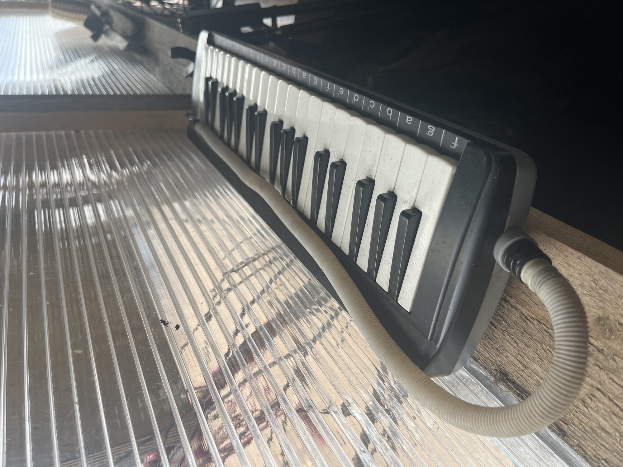 A close-up, angled shot shows a melodica resting on a wooden surface. The instrument is black with white and black keys, and a cream-colored flexible tube extends from its side. The keys are labeled with letters, and the melodica is positioned against a backdrop of translucent, corrugated plastic sheeting. The lighting suggests an indoor environment with natural light filtering through the plastic. The overall scene appears to be a still life of the musical instrument.