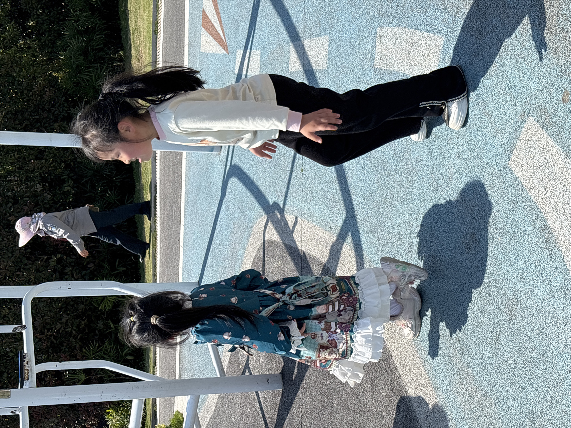 A long outdoor shot shows two children on a light blue, concrete-surfaced playground area bordered by greenery; a third person in a pink hat is partially visible at left. A young girl in a pale-colored sweater, black pants, and white shoes is facing left, balancing on the metal playground bars. Another girl in a dark green dress with a ruffled white petticoat and light-colored shoes is on the ground next to the bars. The scene appears to be in Jiangjin, China.