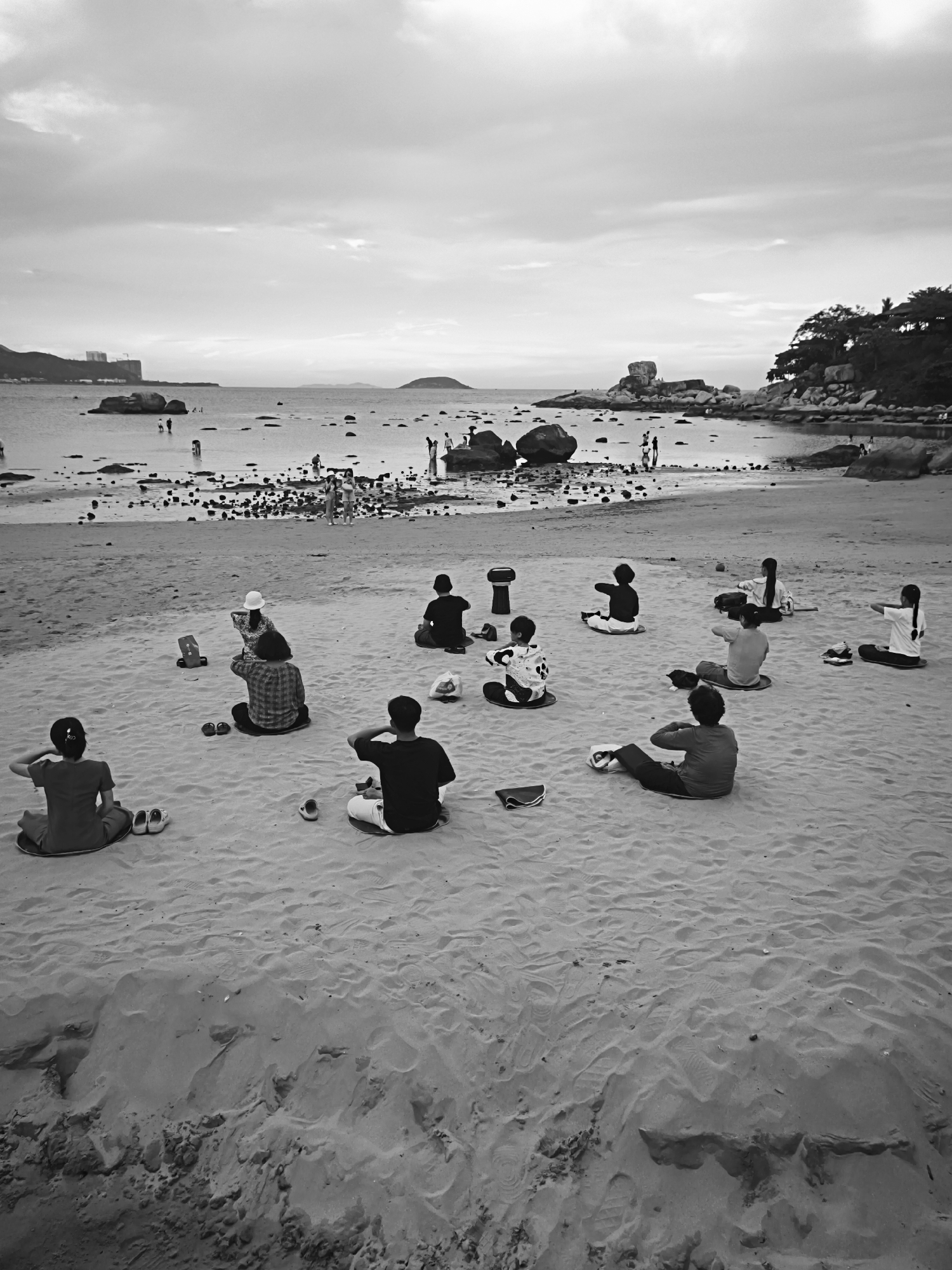 In this black and white photograph, a group of people are practicing yoga on a sandy beach, likely in the city of Bac Nha Trang Ward, Vietnam. The scene is divided between the foreground, where the yoga practitioners are situated, and the background, which depicts the sea and distant land.
In the foreground, approximately ten individuals are seated on the sand in various yoga poses. Most are facing away from the camera, engrossed in their practice. They are arranged in a loose formation, with some individuals closer to the water's edge and others further back. Scattered around them are personal belongings such as shoes, bags, and mats. The sand itself shows the texture of footprints and the uneven surface of a natural beach.
The midground of the image reveals the shoreline, where the sand transitions to shallow water. Several large rocks are scattered throughout this area, and a few individuals are visible wading or standing in the water. Further into the sea, more people can be seen enjoying the water, and some boats or floating objects are present.
The background of the photograph features the expansive sea, with a faint horizon line. On the left side, a few buildings and a hilly landscape are visible, suggesting an urban or semi-urban coastal environment. On the right, a rocky outcrop with dense foliage and some structures on top protrudes into the sea, adding a natural element to the scene. The sky above is overcast, with visible cloud formations, creating a diffused light across the entire image. The overall impression is one of a peaceful, outdoor activity taking place in a scenic coastal location.