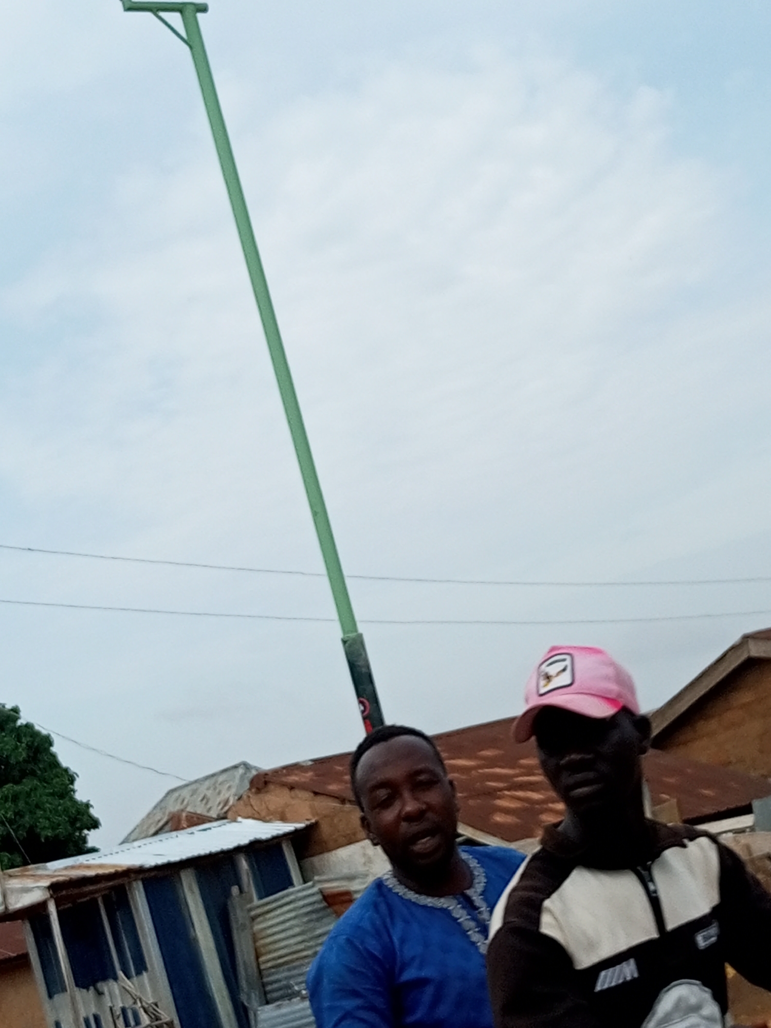 Two men are standing in front of a cluster of buildings under an overcast sky. The man on the left wears a blue garment with white embroidery around the neck. The man on the right wears a pink baseball cap and a black and white jacket. A tall, thin, green pole extends diagonally upward behind the two men. The buildings in the background appear to be made of corrugated metal and possibly mud brick. One building has a white corrugated metal roof, and another has a rusty, corrugated metal roof. A tree with green foliage is visible to the left of the buildings. The scene appears to be in an urban or semi-urban environment in Tumfure, Nigeria.
