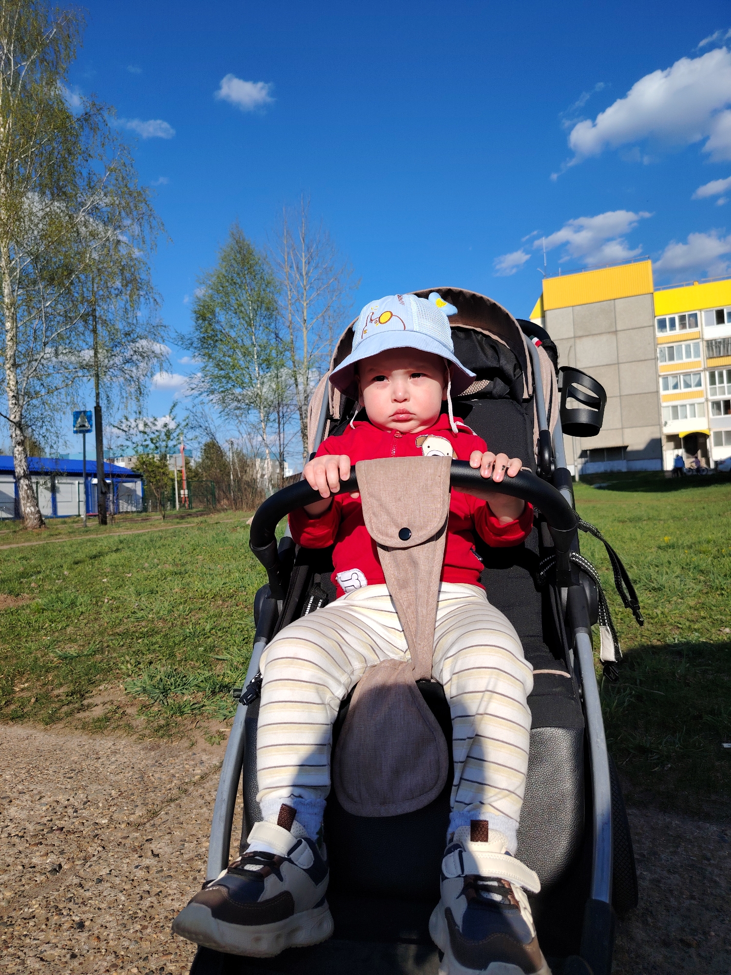 A baby is sitting in a stroller in a park in Марковский, Russia. The baby is wearing a red sweatshirt, striped pants, and a light blue hat. The stroller is black and gray. The background includes grass, trees, buildings, and a blue sky with clouds. A pedestrian crossing sign is visible in the distance.