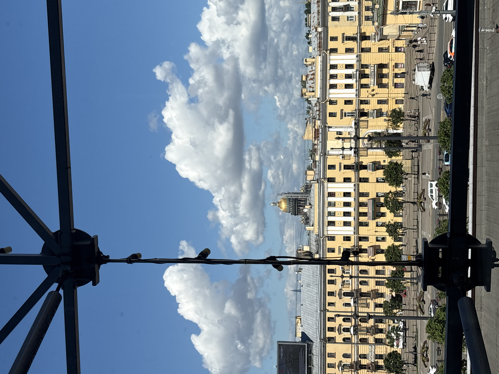 The image is a view of Saint Petersburg, Russia, likely taken from an elevated position, possibly a balcony or rooftop. In the foreground, a dark, metallic structure, possibly part of an awning or canopy, is visible. A thin black cable with what appear to be lights or speakers hangs vertically down the center of the frame.
In the midground, a long, yellow building with multiple floors and numerous windows stretches across the scene. The architecture suggests a historic European style. Trees are planted along the sidewalk in front of this building. Beyond the yellow building, a prominent golden dome of a church or cathedral is visible, silhouetted against a bright blue sky filled with fluffy white clouds.
The street in front of the yellow building shows a moderate amount of activity. Cars are driving on the road, and several people are walking on the sidewalk. There are also some advertisements or signs visible on the building. The overall impression is an urban landscape on a sunny day.