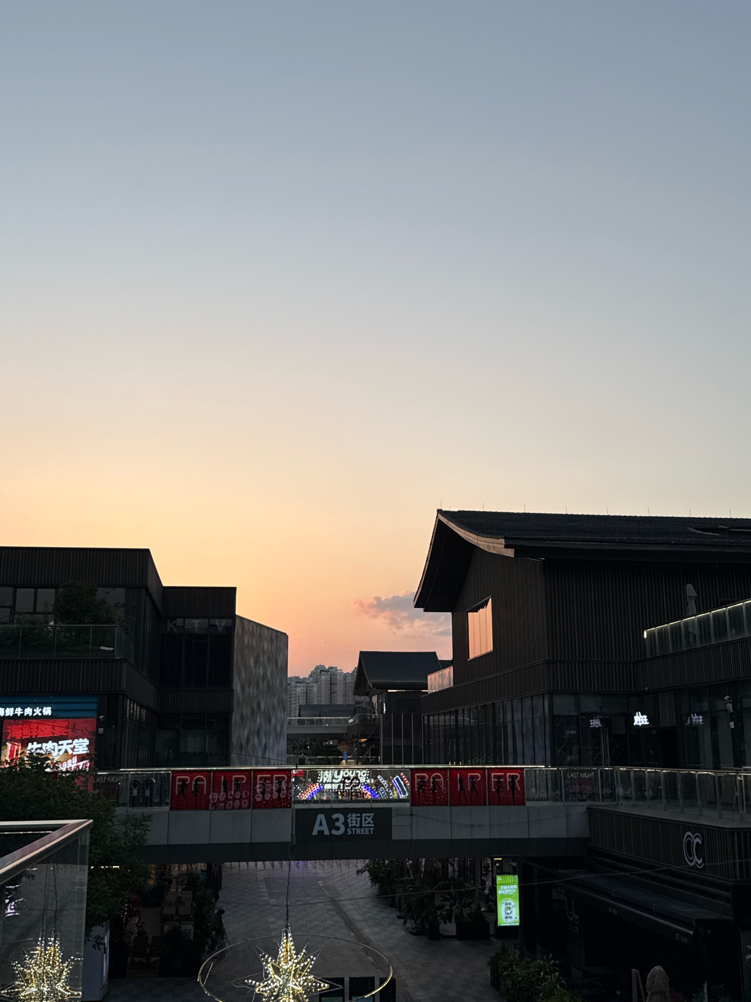 This is a photograph taken at dusk in Yiwu, China, likely at a modern commercial complex. The sky transitions from a pale blue at the top to a warm orange and pink hue near the horizon, indicating sunset. Silhouetted against the sky are several multi-story buildings with dark, contemporary architecture, featuring sharp angles and flat roofs. One building on the right has a prominent pitched roof with a dark wooden exterior.
In the mid-ground, an elevated walkway or bridge spans across an open area, adorned with illuminated signage and decorations. A prominent sign on the bridge reads "A3 STREET". Below the bridge, a pathway is visible, lined with trees and illuminated by decorative star-shaped lights hanging from above. Some of these lights are visible in the foreground. Storefronts with signs are visible on both sides of the pathway.
A large digital billboard on the left side of the image displays text and images, including what appears to be a restaurant advertisement. The overall scene suggests a bustling urban environment, possibly a shopping district or entertainment area, at the end of the day. No people are clearly discernible in the image, but the presence of commercial establishments and decorative lighting implies human activity.