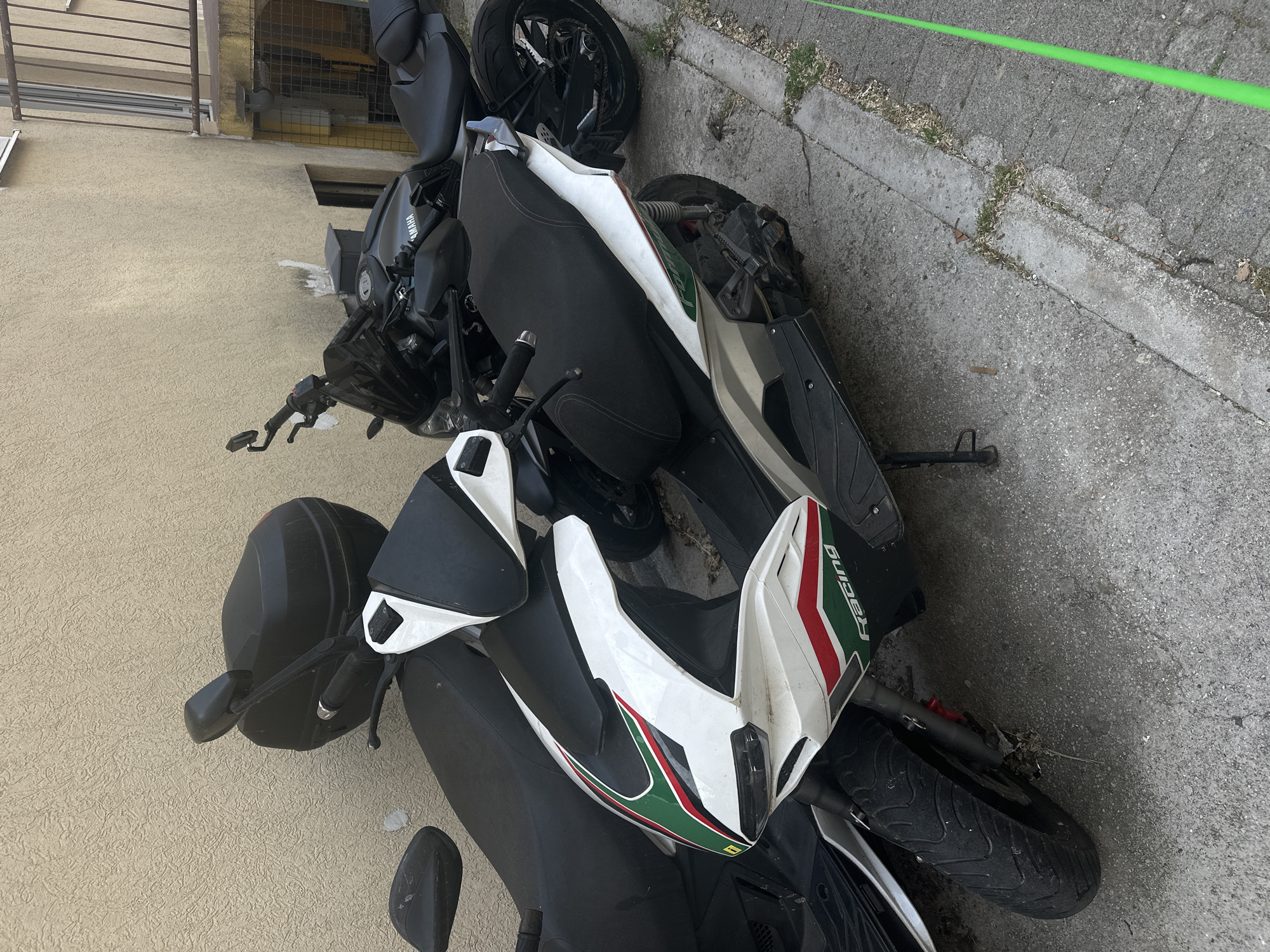 Two motorcycles, a black Yamaha and a white and black Racing scooter with Italian-flag-colored stripes, lie horizontally on the ground. One rests against a pale yellow wall and the other on concrete with a green rope or hose running along its edge. The scene appears to be outdoors. The location is potentially Budva, Montenegro.