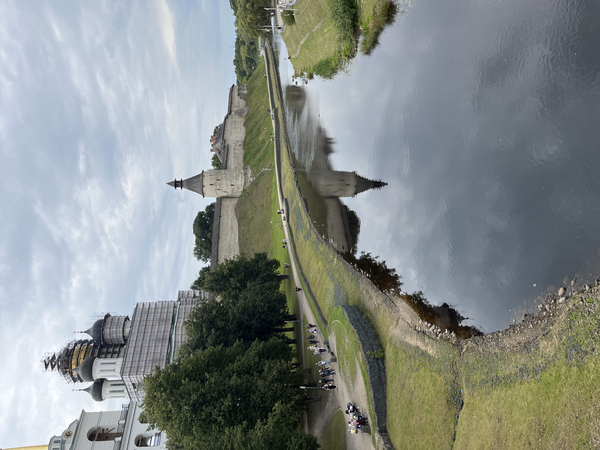 The image captures a view of the Pskov Krom (Kremlin) in Pskov, Russia, from across a river. On the left, a white church with several domes is partially covered in scaffolding, indicating ongoing renovation work. The church has a golden spire visible at the top. Adjacent to the church, a stone fortress wall with a tower extends along the riverbank. Several people are walking along a paved path on the grass-covered embankment in front of the fortress. The river in the foreground reflects the sky and the structures on the opposite bank. The sky is overcast with clouds. The overall scene depicts a historical landmark with signs of preservation work and public activity.