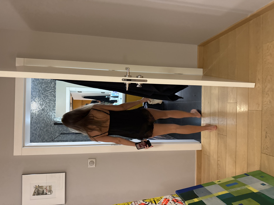 A woman in a black nightgown stands in a doorway, facing away from the camera. She holds a phone in her left hand. The door is open, revealing a bathroom with mosaic tiles and a glimpse of another room beyond. The hallway features light wood flooring and a gray wall adorned with framed artwork. Part of a colorful abstract painting is visible in the foreground. A black garment hangs on the open door.