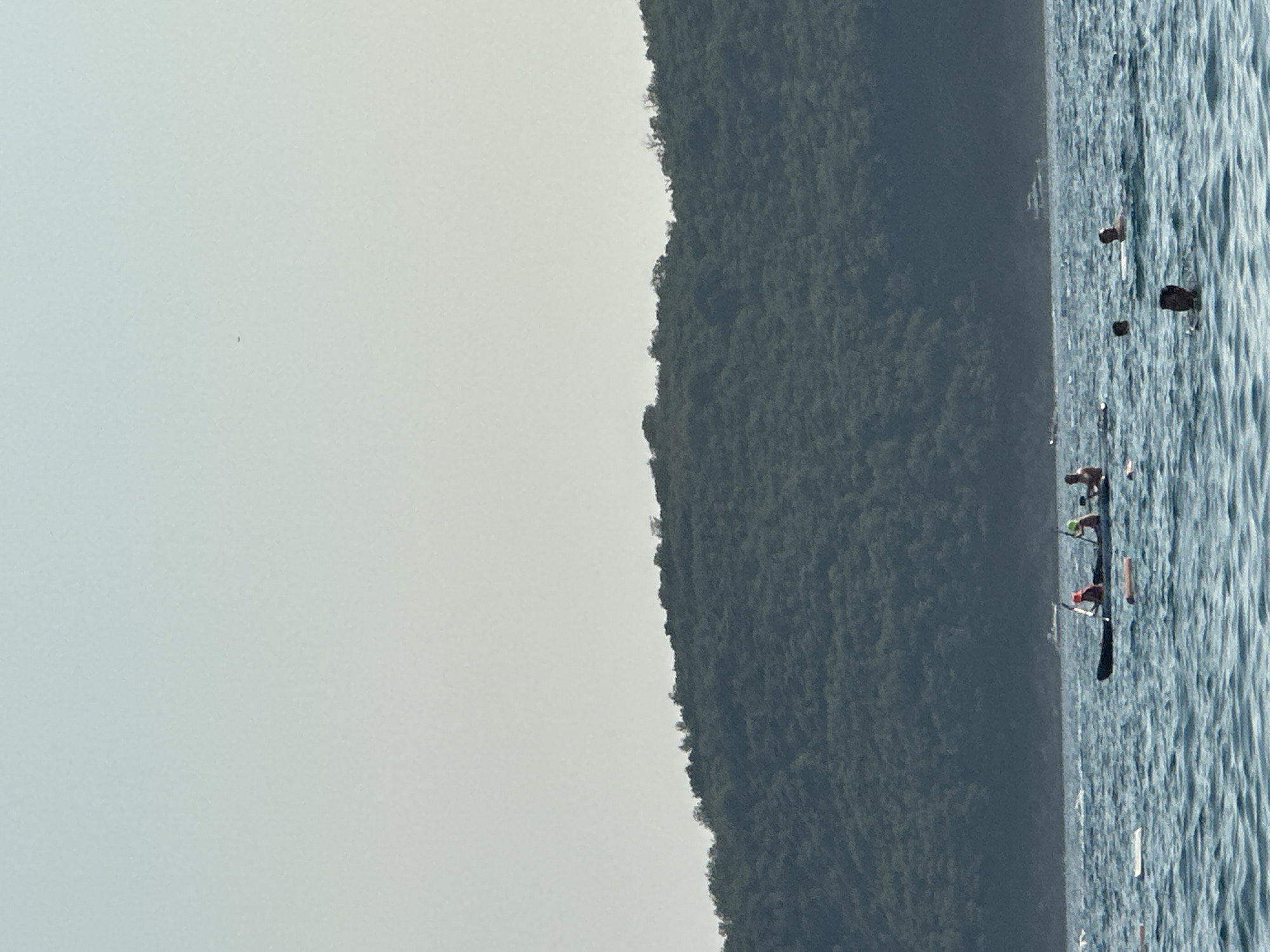 The image depicts a serene coastal scene, likely in Boreti, Montenegro, characterized by a tranquil sea and a verdant, forested hill. The water is a deep blue, with gentle ripples indicating calm conditions. Several individuals are visible in the water, some swimming and others on flotation devices. In the middle ground, a kayak carries three people, two of whom are wearing life vests, suggesting recreational activity. Further in the distance, a white boat is anchored, its presence adding a touch of activity to the otherwise peaceful seascape. The backdrop is dominated by a lush, green hill that slopes down towards the water's edge, its dense foliage creating a natural and picturesque setting. The sky is a pale, hazy blue, suggesting fair weather.