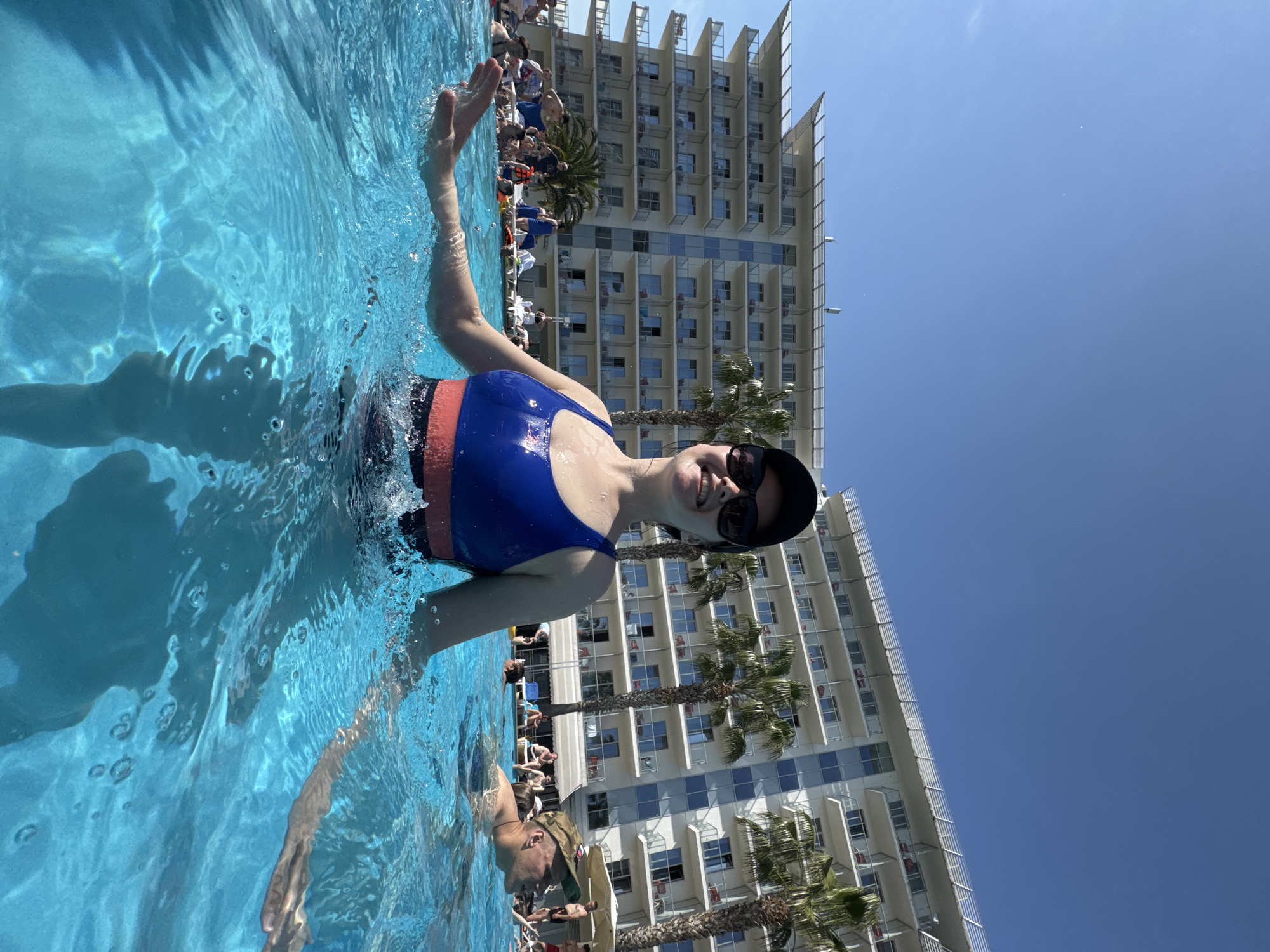 The photograph captures a woman in a swimming pool, likely at a resort given the backdrop of a tall building and palm trees. The woman is wearing a blue and coral-colored swimsuit, sunglasses, and a hat. She is smiling and waving to the camera. The pool water is clear and blue, and there are other people visible in the background, both in the pool and lounging on chairs. The building behind her is tall and appears to have multiple balconies. The sky is a clear, bright blue.
Due to the lack of recognizable landmarks, architectural styles, or unique environmental features specific to Sochi, it is not possible to definitively confirm that the image was taken in Sochi, Russia.
