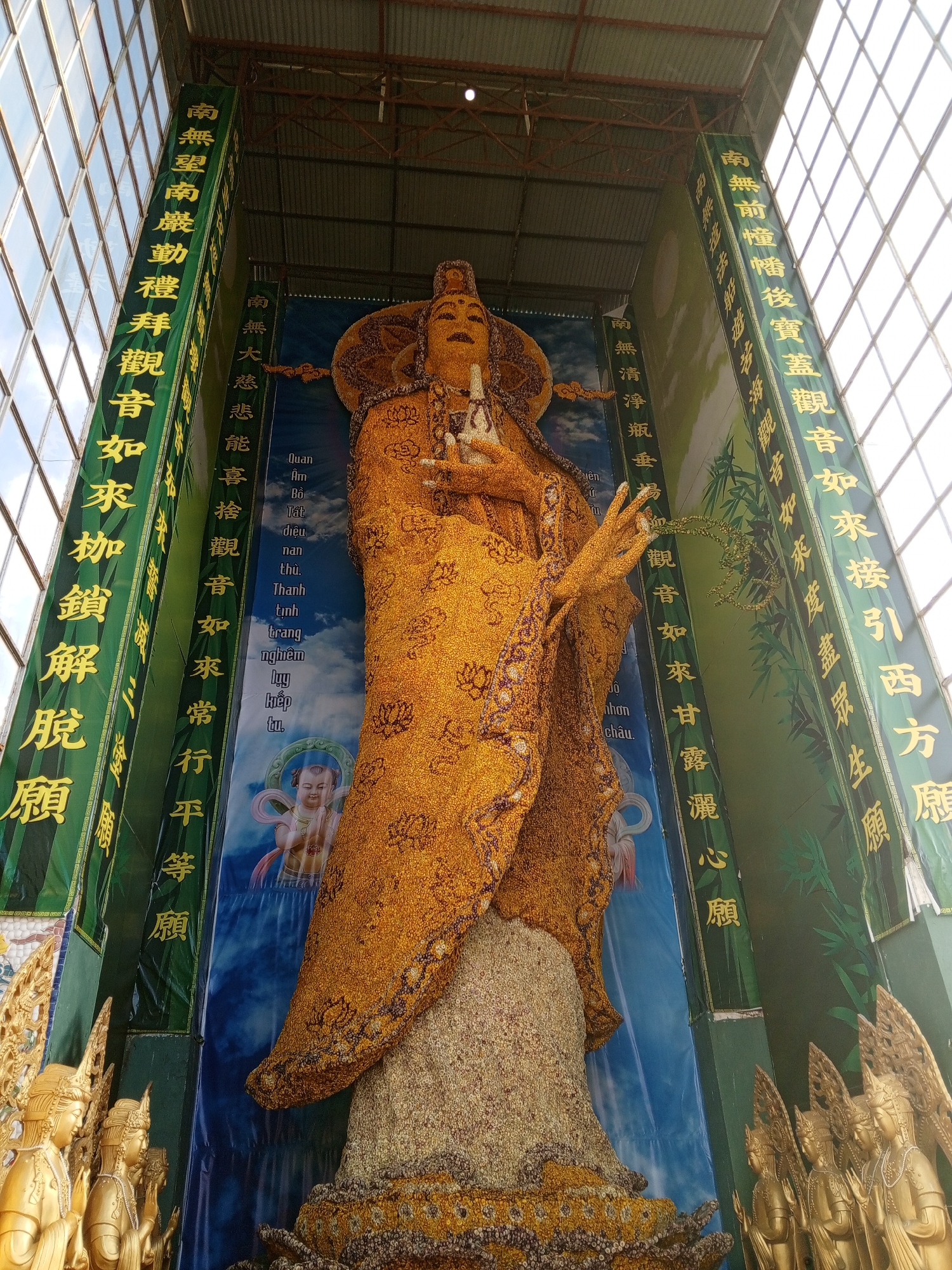 In the city of Xuân Thành, Vietnam, a massive, gilded statue of a Buddhist deity, likely Guanyin, stands prominently within an open-air structure. The statue is adorned with what appears to be a dense covering of small, golden-yellow flowers, giving it a textured and vibrant appearance. It is framed by tall, green banners featuring golden Chinese characters, which are likely Buddhist prayers or dedications. These banners are hung vertically on either side of the statue, creating a visual corridor.
The background behind the statue is a large blue banner with a depiction of clouds and a smaller, serene figure of a child-like deity, possibly also Guanyin in a different manifestation. To the left and right of the main statue, at its base, are smaller golden statues of what appear to be Buddhist attendants or devotees, depicted with hands pressed together in prayer. The entire scene is illuminated by natural light filtering through a large, grid-patterned glass ceiling or windows above, suggesting the structure is a temple or a dedicated sanctuary. The ceiling structure itself is made of exposed metal beams, indicating an industrial or functional construction.