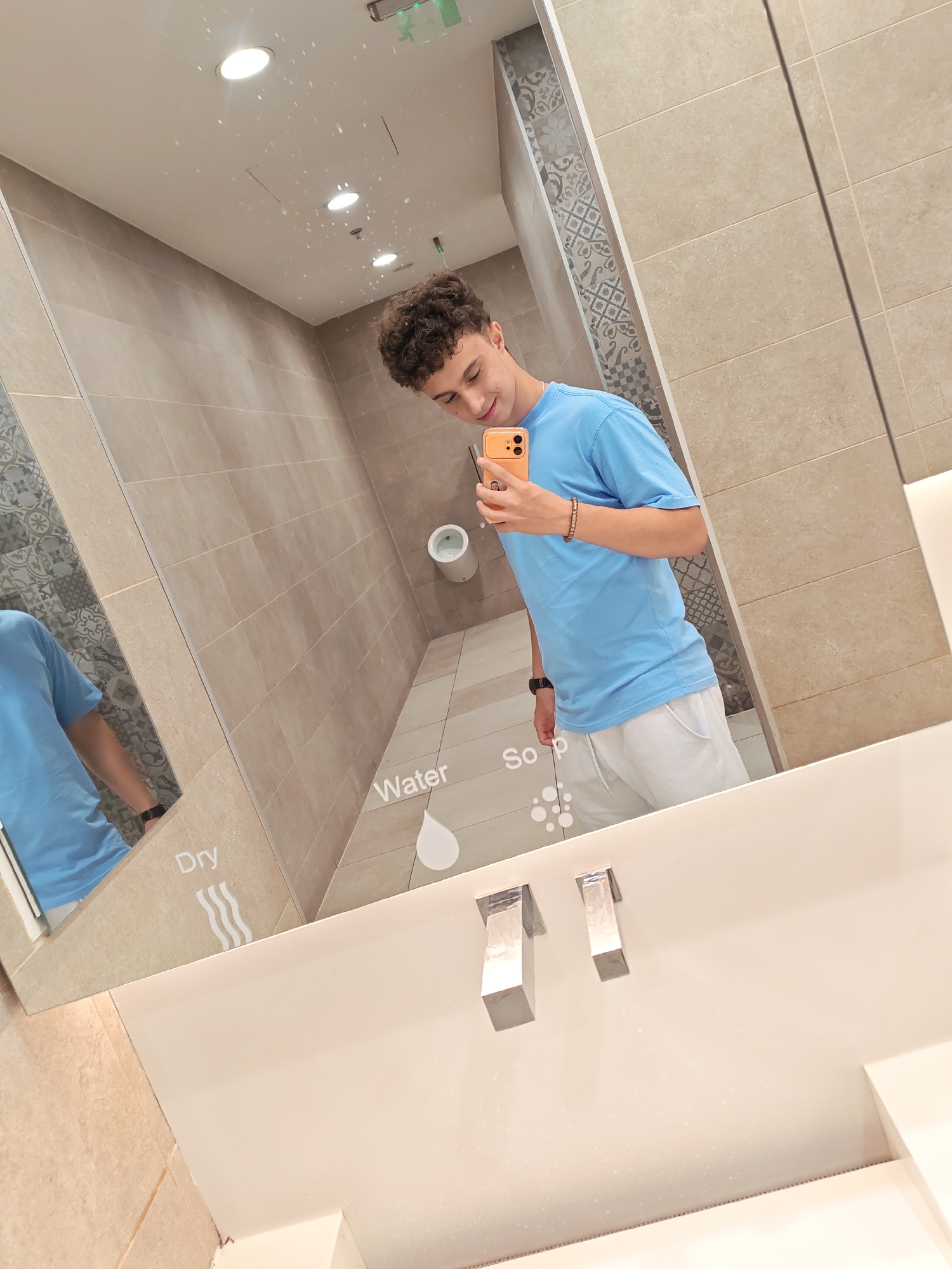 A young male with curly hair is taking a selfie in a modern restroom. He is wearing a blue t-shirt and white shorts, and holding an orange smartphone. The restroom features beige tiled walls, a white sink with two chrome faucets, and a large mirror reflecting the scene. Text and icons indicating "Dry," "Water," and "Soap" are visible on the mirror's surface. The overall impression is one of a clean and contemporary public space. The location is identified as Stara Zagora, Bulgaria.