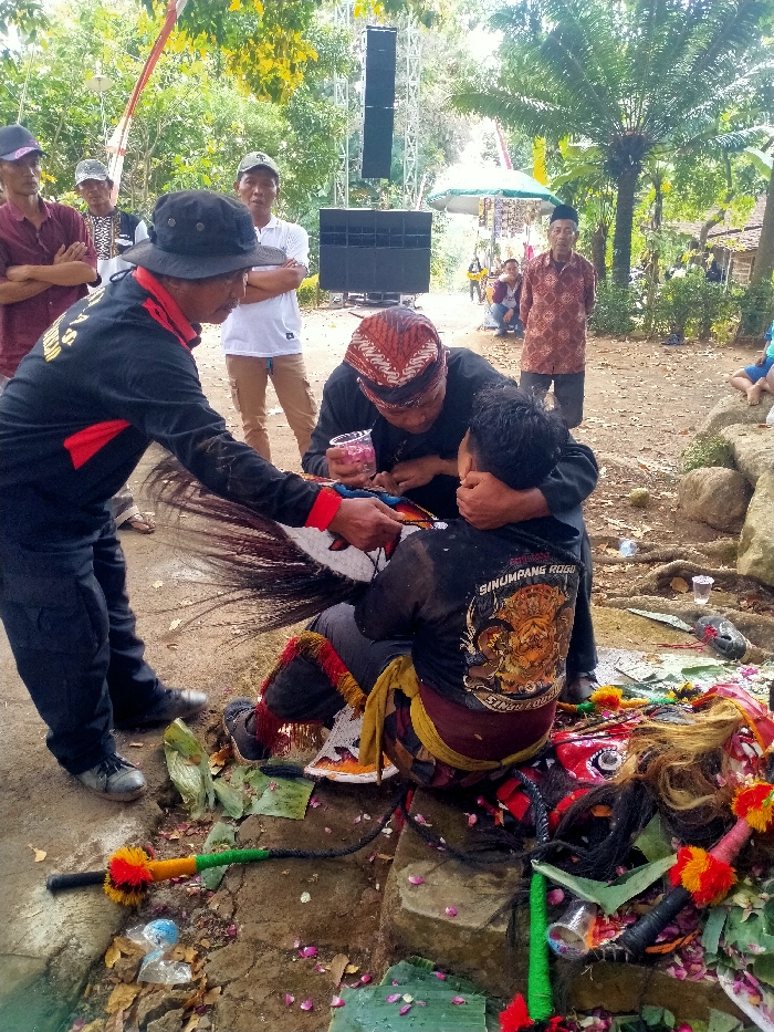 The image depicts an outdoor scene in Menjing, Indonesia, featuring a traditional event or ritual. In the immediate foreground, three adult males are engaged in a central activity on an unpaved dirt ground marked by scattered green banana leaf pieces and red/pink flower petals.
To the left, a standing male, attired in a black long-sleeved shirt with red accents, black trousers, and a dark boonie hat, leans forward, extending his right hand towards a white and red stylized horse prop (kuda lumping) with a prominent black tail.
In the center, a male wearing a black shirt and a traditional reddish-brown batik-patterned headwrap is kneeling or bent over. He holds a clear plastic cup containing red and pink flower petals in his left hand and has his right arm around the shoulder of the third male. He appears to be performing a ritualistic action or preparation.
To the right, a male, wearing a black long-sleeved shirt with a yellow, orange, and red "SINUMPANG ROGO" graphic on the back, is seated or crouched low. He is adorned with patterned traditional leg coverings and has his left hand covering his mouth/chin area.
On the ground surrounding these individuals are various objects consistent with traditional performances: a multi-colored whip-like object (pecut), and at least two traditional red and black masks or headdresses, laid on banana leaves and surrounded by petals. Several clear plastic cups and miscellaneous plastic debris are also present.
In the background, a large sound system, consisting of a tall metal structure holding multiple black line array speakers and a subwoofer, is prominently displayed. Several male spectators are observing the scene, some with crossed arms. The environment is characterized by dense green trees and foliage, including palm trees, indicating a natural or semi-rural setting. The ground is primarily unpaved earth with some visible concrete or stone edging.