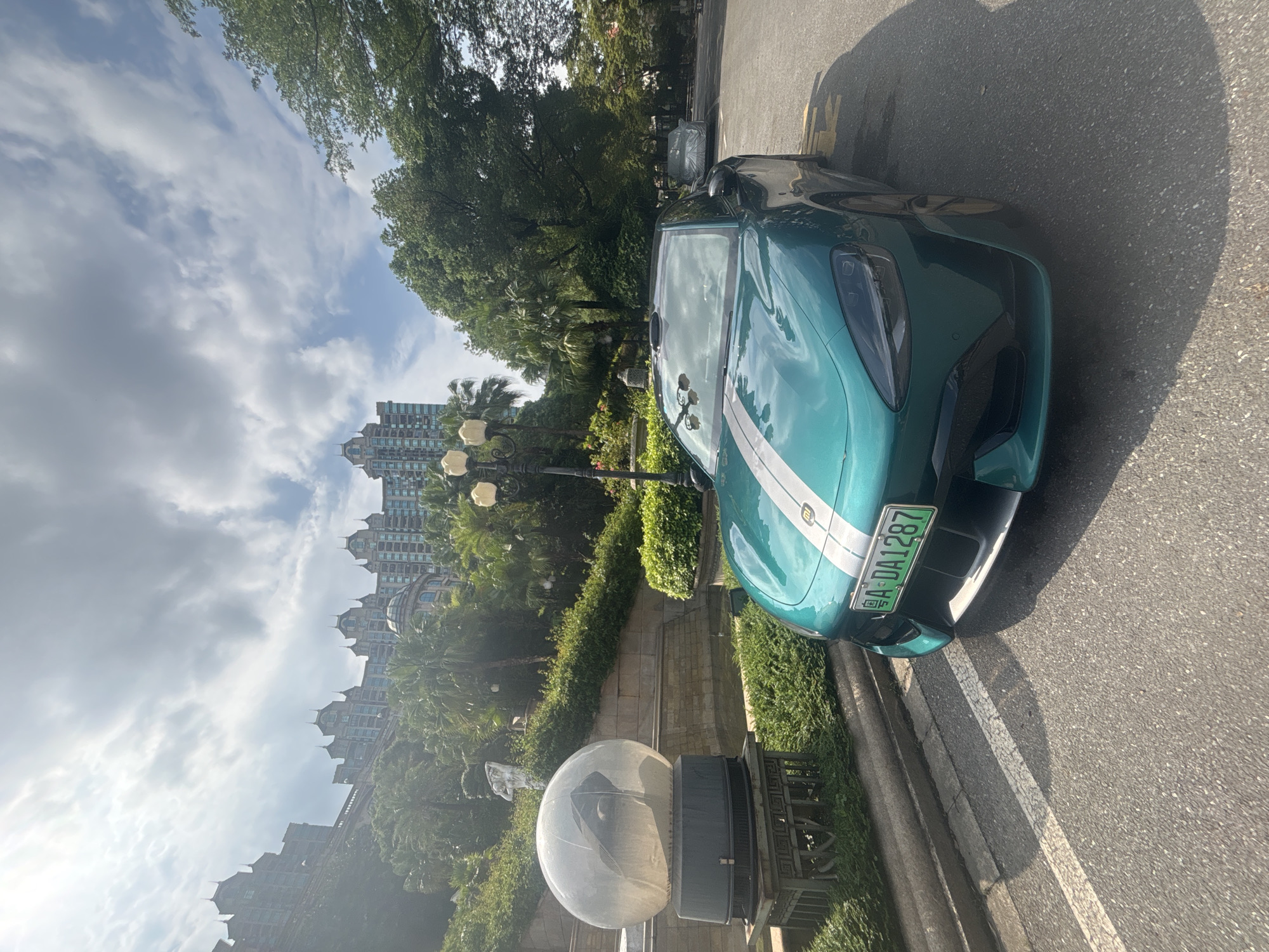 A teal-colored electric car, identified as a Xiaomi SU7, is parked on an asphalt road in front of lush greenery and a large, ornate building complex that appears to be a resort or hotel. The car has white racing stripes running down the hood and roof. Its license plate reads "A DA1287". In the background, the building features numerous domes and intricate architectural details, resembling a castle or palace, with palm trees and other tropical foliage surrounding it. To the left of the car, a large, clear spherical sculpture sits on a pedestal. The sky is partly cloudy. The scene appears to be a sunny day in a well-maintained, upscale area, likely a tourist destination in the Nanhai District of China.