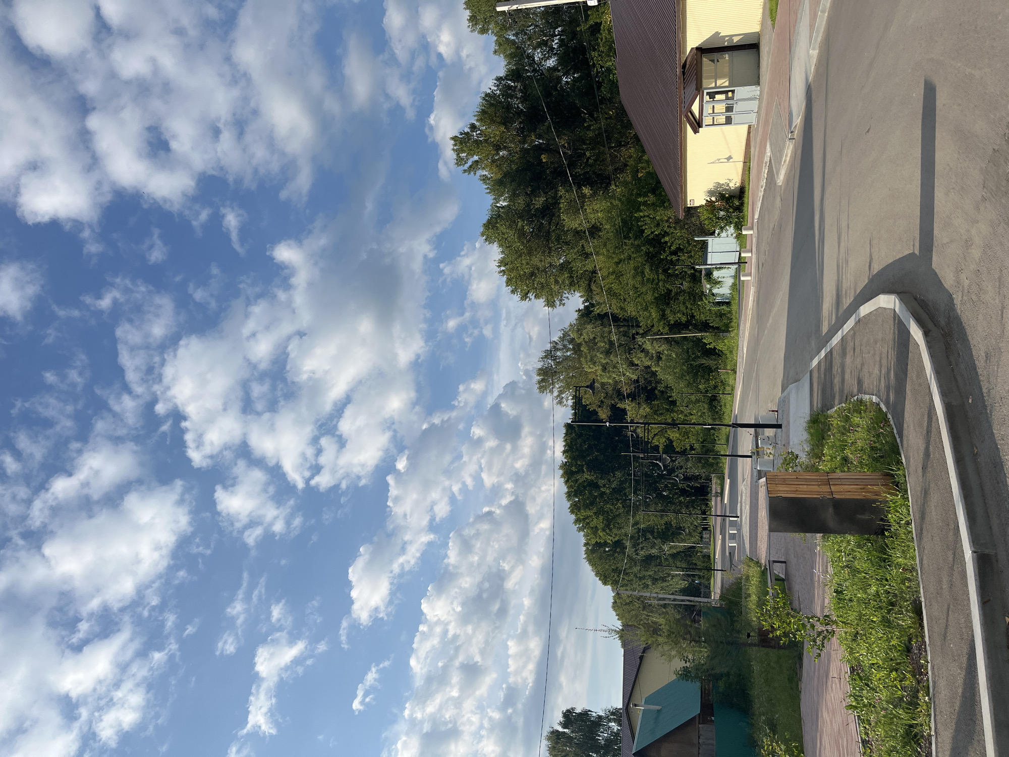 The image depicts a paved road in a residential area, likely in Blagoveshchensk, Russia. The road curves to the right in the foreground, bordered by a sidewalk and grassy areas with landscaping. On the left side of the road, there's a building with a green roof and a fence, followed by more greenery and trees. On the right side, a yellow building with a brown roof and a covered entrance is visible. The background is dominated by a dense line of trees. The sky is a vibrant blue with scattered white, fluffy clouds, suggesting a clear day. There are no people visible in the image, and no actions or interactions are taking place. The scene appears to be a quiet, recently developed or maintained suburban street.