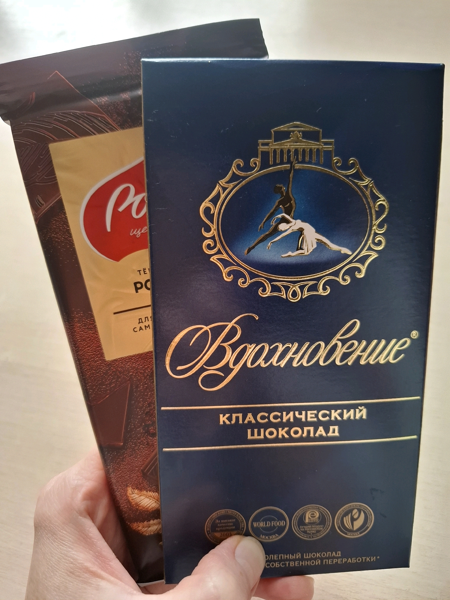 The image shows two chocolate bars being held by a person's hands. The chocolate bar in the foreground is dark blue with gold lettering that reads "Вдохновение" (Inspiration) and "КЛАССИЧЕСКИЙ ШОКОЛАД" (Classic Chocolate). It also features a golden emblem with silhouettes of two dancers. The chocolate bar behind it is dark brown with a golden background and red lettering that says "PO" (a partial view of the brand name). The scene is indoors, with a light-colored wooden surface visible in the background, suggesting a table. The image does not contain any information that would allow for the identification of the city of Tyumen, Russia, or any specific urban environment.