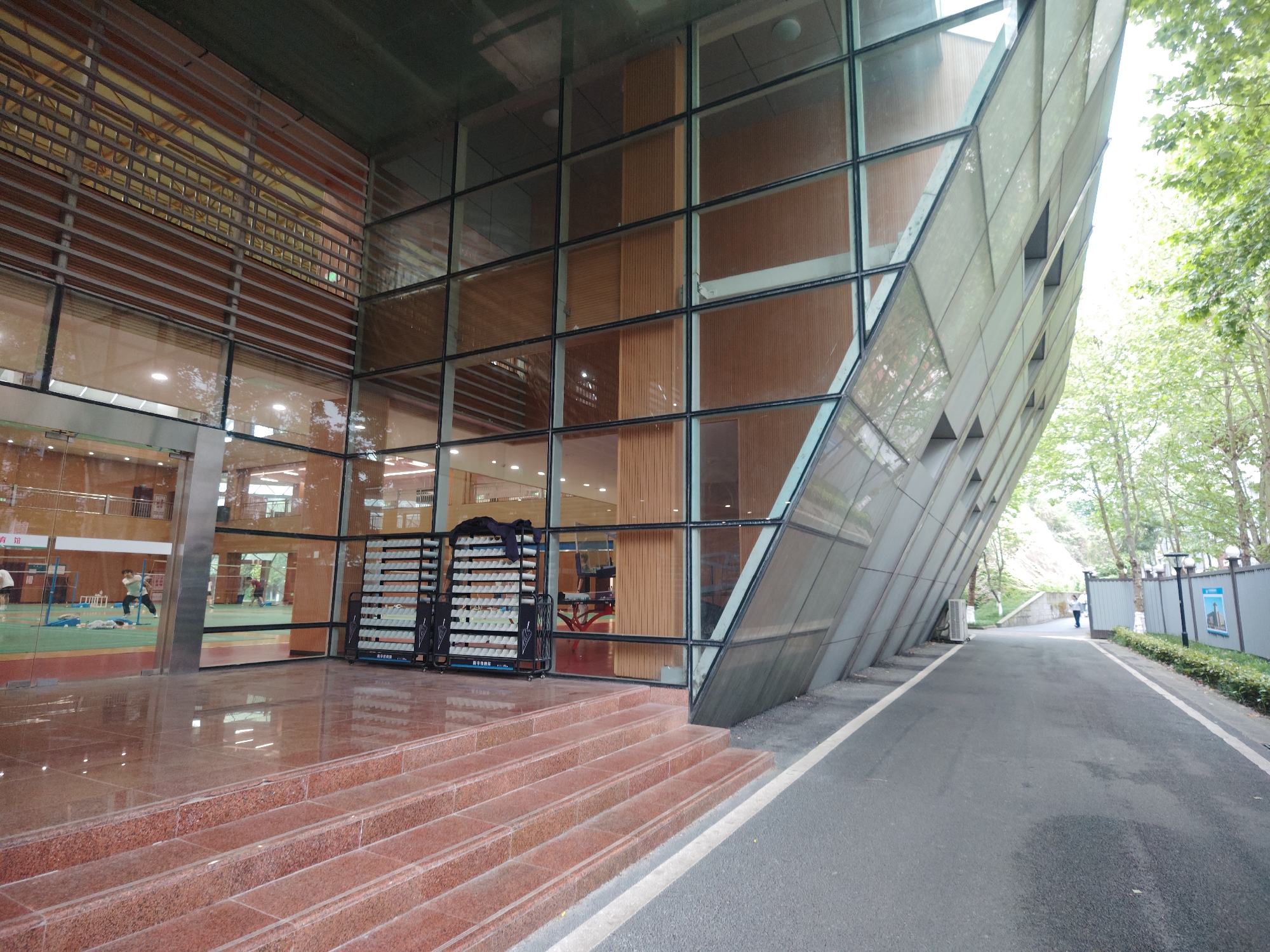 The image captures the exterior of a modern building in Chongqing High-tech Zone, China. The building has a unique architectural design with a large glass facade structured into a grid pattern, with sections of the facade angled outwards. The building's interior is visible through the glass, revealing a gymnasium with people engaged in activities. Outside the building, there are stairs leading up to the entrance, and two shoe racks are placed near the steps. The pavement runs alongside the building, leading towards a road with a pedestrian and a wall or fence with lampposts and greenery. The environment includes trees and hints of a hill in the background.