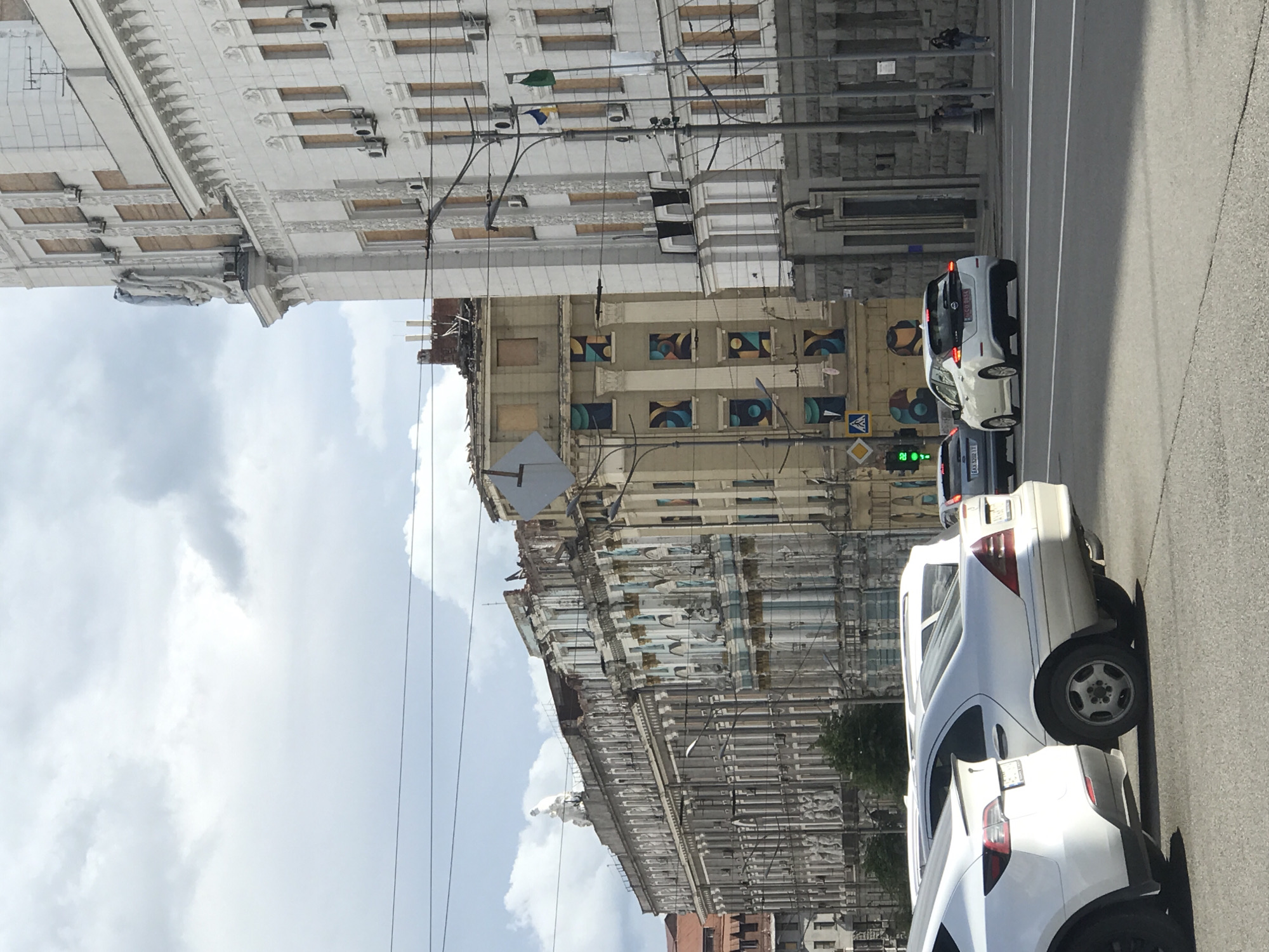 The image depicts a street scene in Kharkiv, Ukraine. Several vehicles, mostly white passenger cars, are stationary in the right side of the road, lined up vertically in the frame. A traffic light showing green is visible. Buildings line both sides of the street, which appear old and weathered. The buildings have decorative facades and appear to be in varying states of repair. There is a building undergoing renovation with painted decorations on its facade. The sky is visible above the buildings, featuring a cloudy atmosphere. Utility wires are strung across the street. The image orientation is rotated 90 degrees counterclockwise.