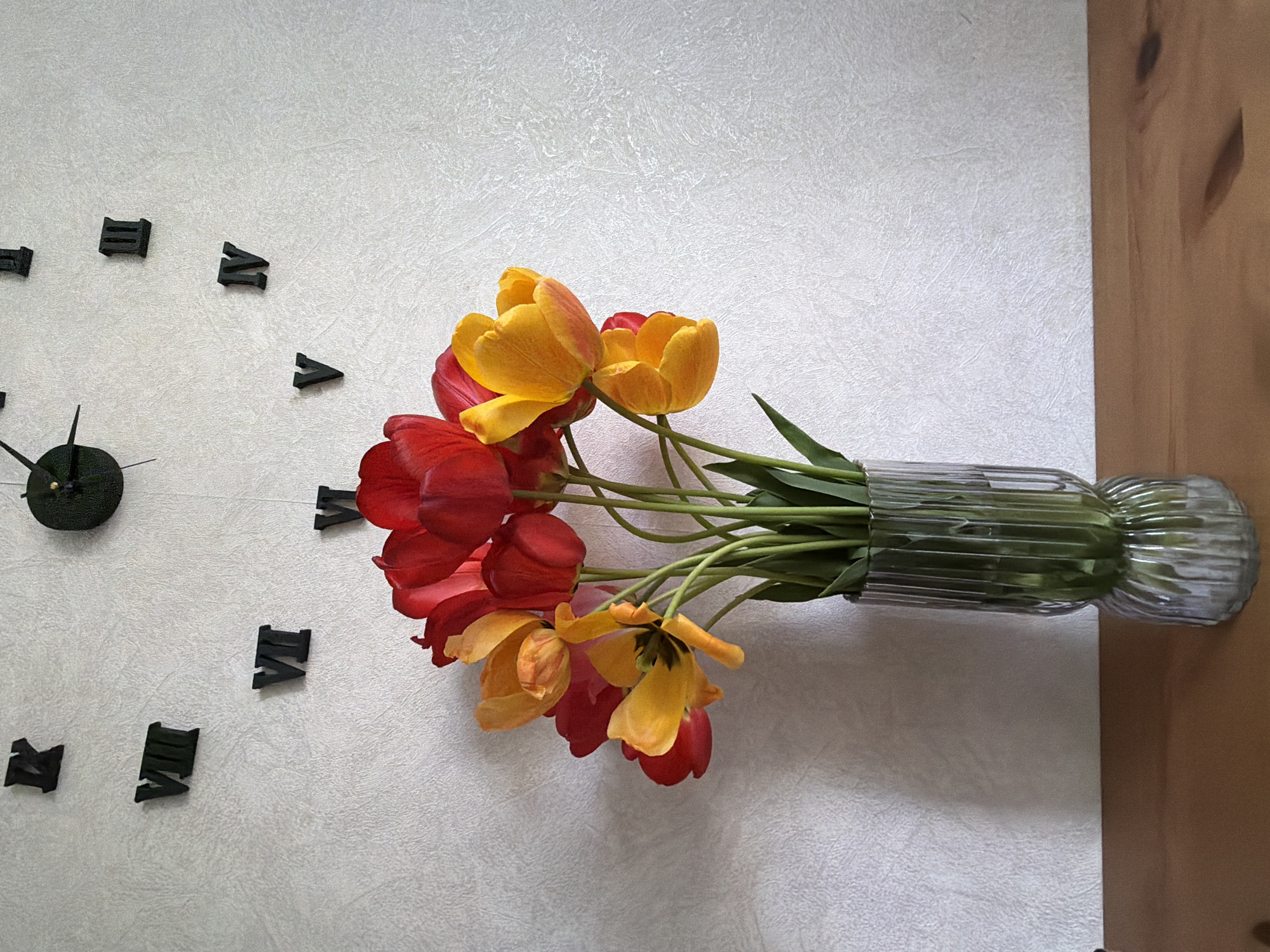The image shows a top-down view of a bouquet of red and yellow tulips in a fluted glass vase. The vase rests on a white surface with a slightly textured pattern. A portion of a wooden surface is visible on the right edge. To the left, a clock is present, indicated by Roman numerals and clock hands attached to a central point. The time indicated is approximately 9:20. There is no evidence to confirm that the location is Tokkari, Russia.