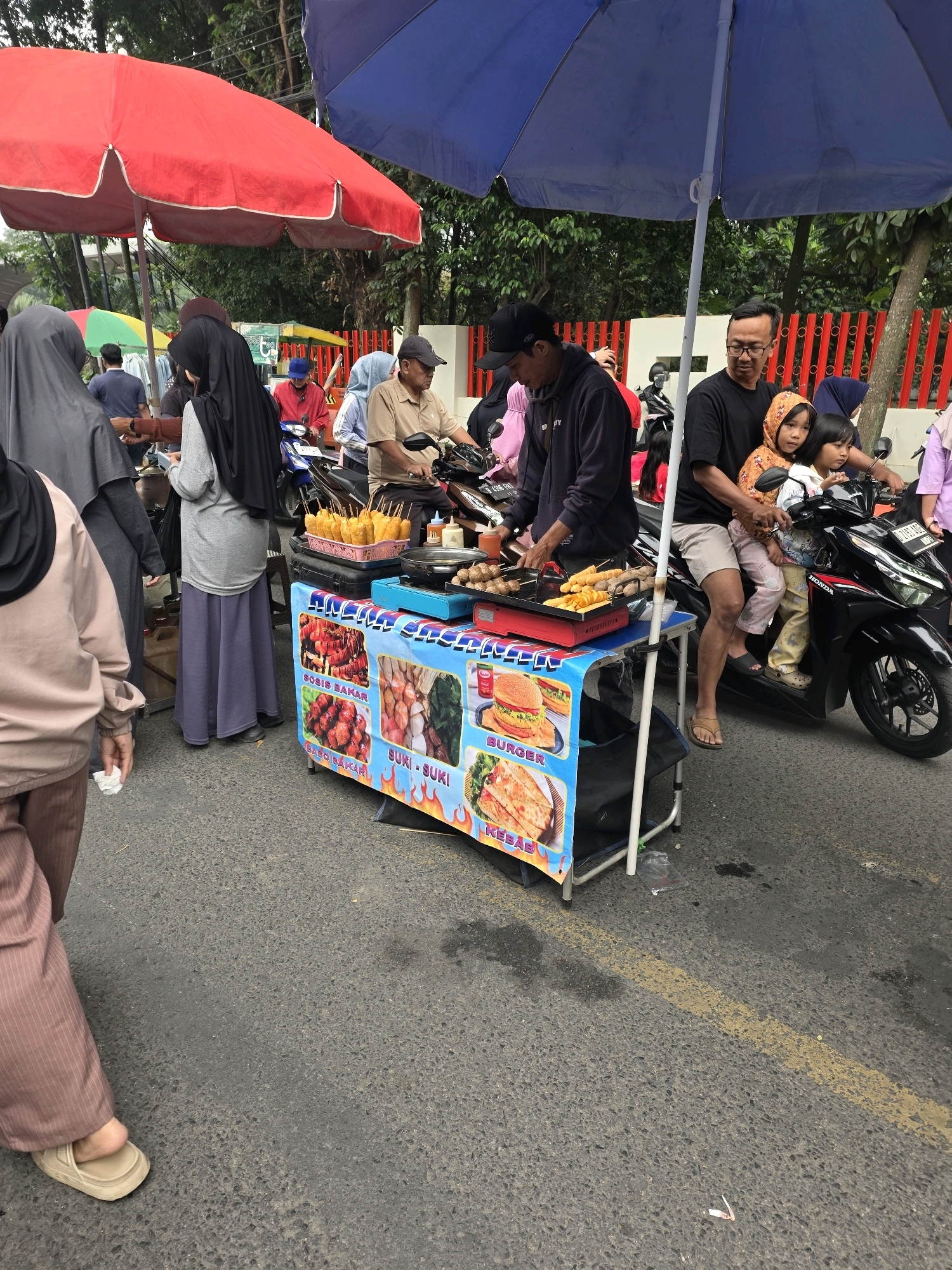 The image depicts a street scene in Bojongsoang, Indonesia, likely near a busy road given the presence of a yellow line on the asphalt. The primary focus is a food stall selling grilled items and burgers, characterized by a colorful banner displaying images of food such as kebabs and sausages. Underneath a blue umbrella, a vendor is preparing food on a grill, with skewers of food visible. In front of the stall, a man is seated on a motorcycle with two young girls, one of whom appears to be a child and the other an infant. They are all looking towards the food stall.
Other individuals are present in the background, including a woman in a black hijab standing near the stall and another person, partially visible, wearing a beige long-sleeved shirt and a headscarf. A woman in a brown outfit with pinstripes is walking past the food stall. Red and blue umbrellas are set up, suggesting a temporary market or roadside dining area. The environment appears to be outdoors with trees and a red fence visible in the background. The overall atmosphere suggests a casual, possibly commercial setting with people engaged in everyday activities.