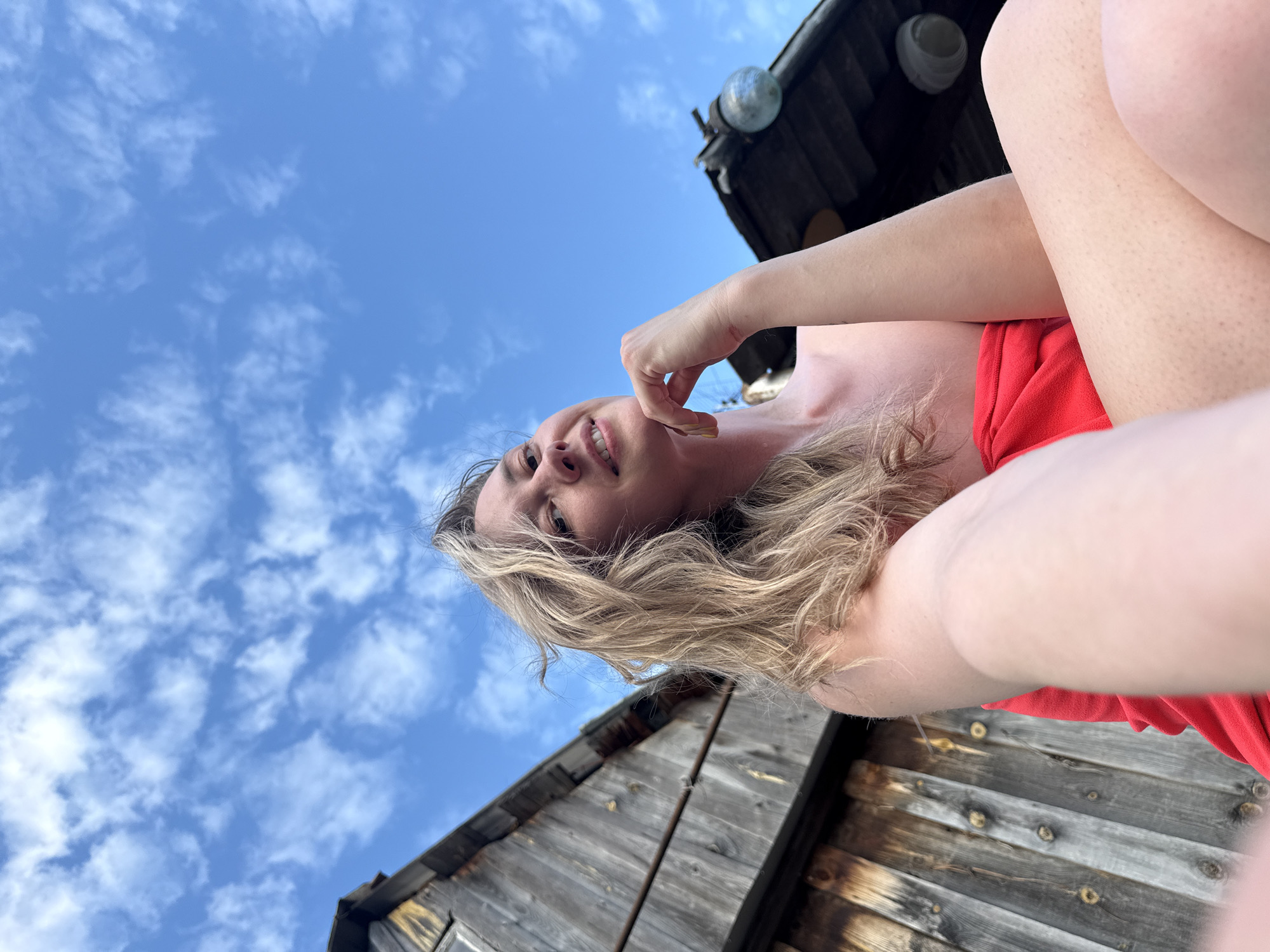 A low-angle, selfie-style photograph shows a woman with blonde hair, wearing a red top, against a backdrop of a blue sky with scattered white clouds. She is touching her chin with her right hand and smiling slightly. The surrounding environment appears to be a wooden structure, possibly a building or a platform. The photograph was taken from an unusual perspective, causing the subjects to appear tilted relative to the horizon. The location is possibly in the city of Chaikovsky, Russia.