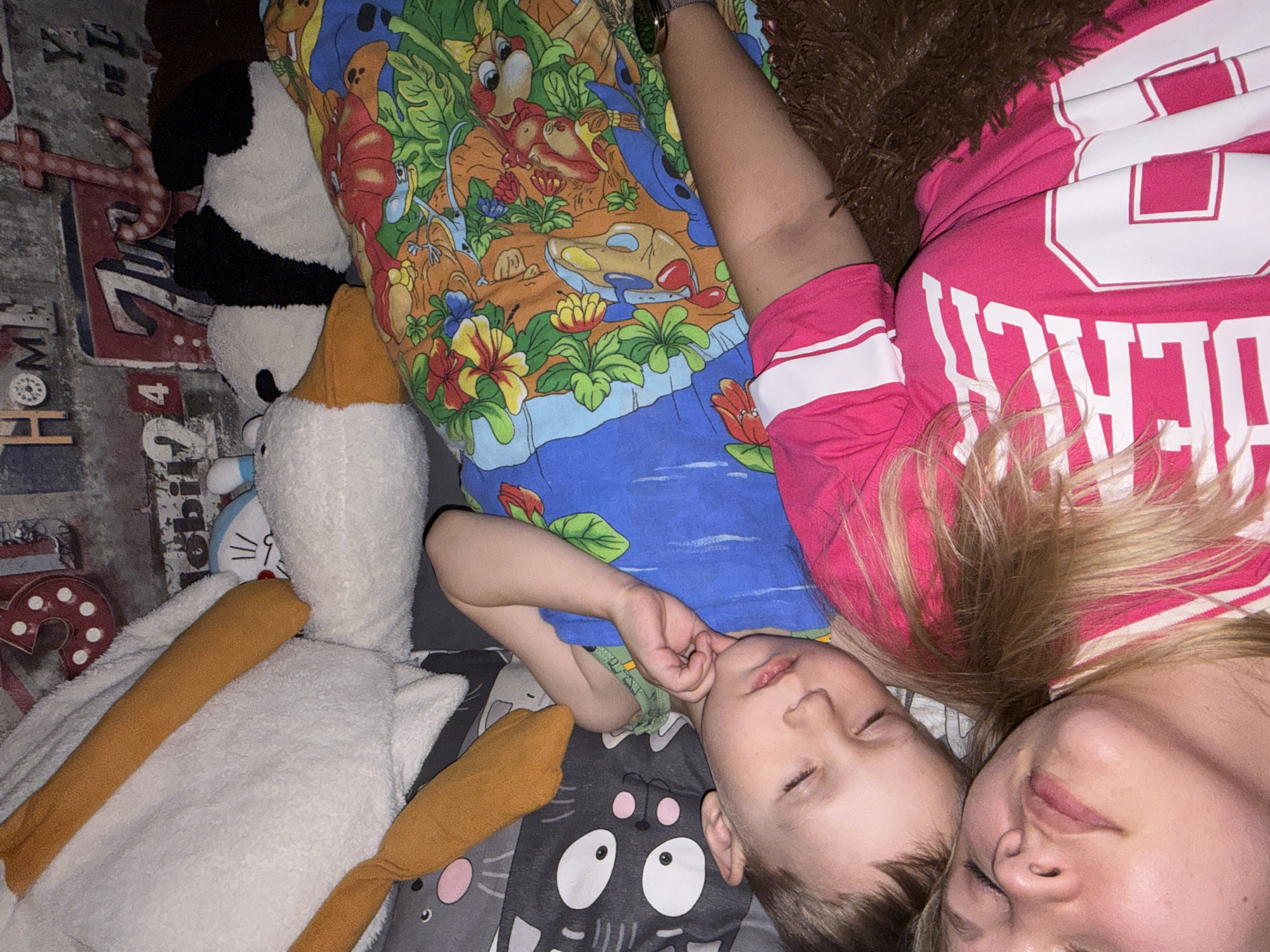 A woman and a young child are lying down, likely sleeping, on a bed or floor. The woman is on the right, wearing a pink shirt with white stripes and lettering. The child is to her left, wearing a blue top with colorful cartoon-like characters and floral patterns. There are several stuffed animals around them, including a large white duck with orange legs and a panda bear. The bedding appears to be patterned with cartoon cat faces. The location is Chaikovsky, Russia.