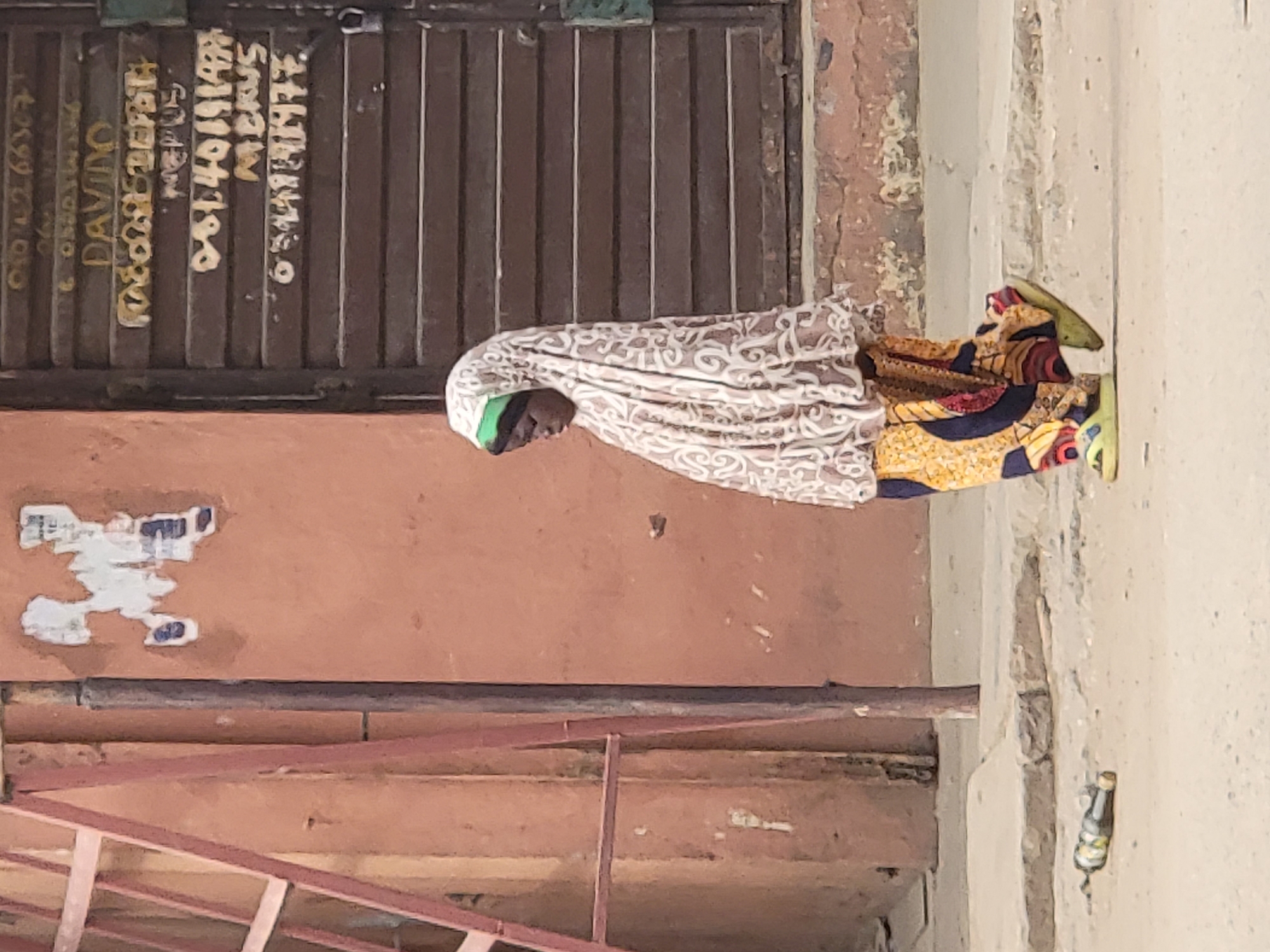 A woman is seen on a city sidewalk in Maiburiji, Nigeria, near a building with a rusted metal door and faded posters on the side. She is wearing a patterned headscarf and a colorful dress, and is stooped over. A discarded bottle lies nearby on the sidewalk.