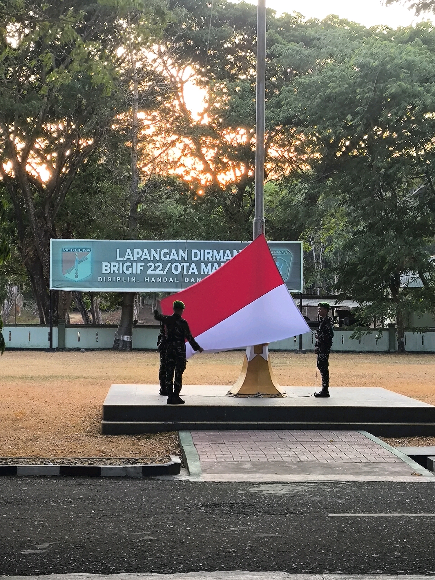 The image depicts an outdoor scene in front of a building, likely a military facility, based on the presence of soldiers and a banner indicating a military unit. The setting appears to be a parade ground or open field, with a flag pole at the center. Two soldiers in camouflage uniforms are standing near the base of the flag pole, holding a large Indonesian flag, which is red and white. One soldier is actively engaged in handling the flag, while the other stands at attention. A banner with text and a crest is visible in the background, suggesting the name of the military unit is "BRIGIF 22/OTA MANASA." The environment is characterized by trees and vegetation, with sunlight filtering through the leaves, indicating it might be early morning or late afternoon. The ground is covered with sand and grass, and there are some paved areas and structures in the foreground and background. The overall impression is a formal ceremony or flag-raising event.