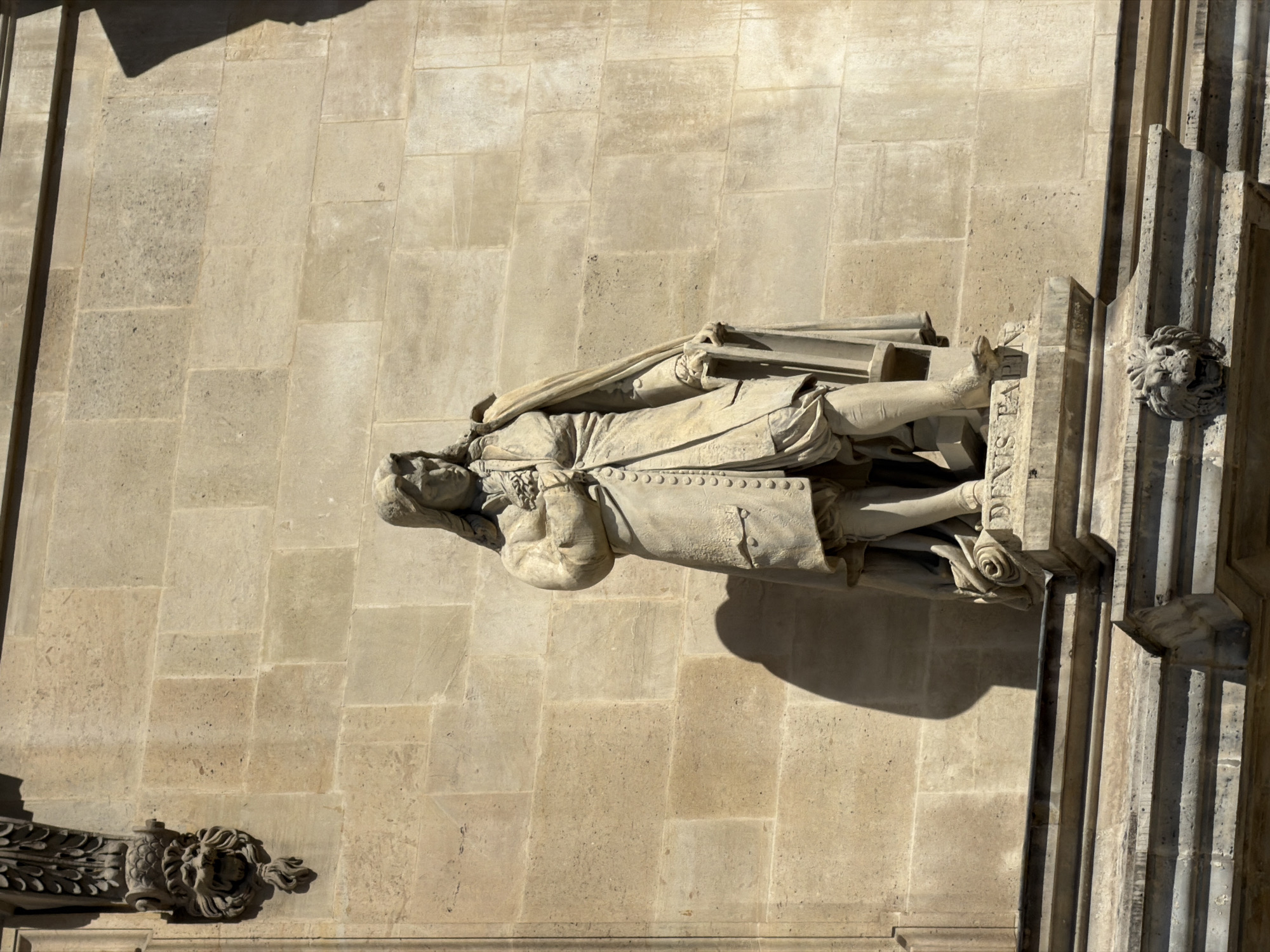 A statue of a man is affixed to a stone building in Paris, France. The man is dressed in 17th or 18th century garb. A stone wall is in the background. A stone ornament is on the bottom left. A pillar is visible in the right foreground.