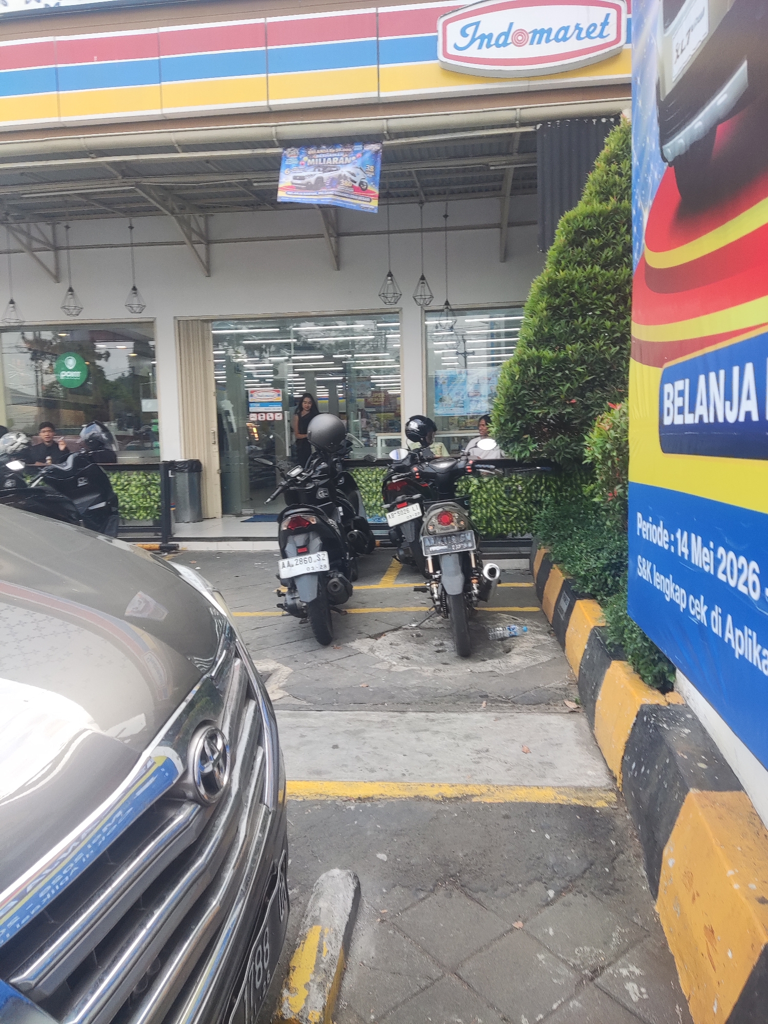 This image was taken in Trihanggo, Indonesia. It shows the exterior of an Indomaret convenience store. In the foreground, the hood and grille of a silver car are visible. To the right of the car, a black motorcycle with a license plate starting with "AA" is parked. Further back, more motorcycles are parked in front of the store. A tall, dense green hedge is located between the store and a blue and yellow advertisement banner. The store has a white awning with a red, blue, and yellow striped border. A sign with the Indomaret logo is displayed prominently above the entrance. Inside the store, displays and shelves are visible through the glass windows. The ground in front of the store is paved with concrete tiles and yellow and black striped concrete barriers line the parking area. A person, likely a woman, is standing inside the store near the entrance.