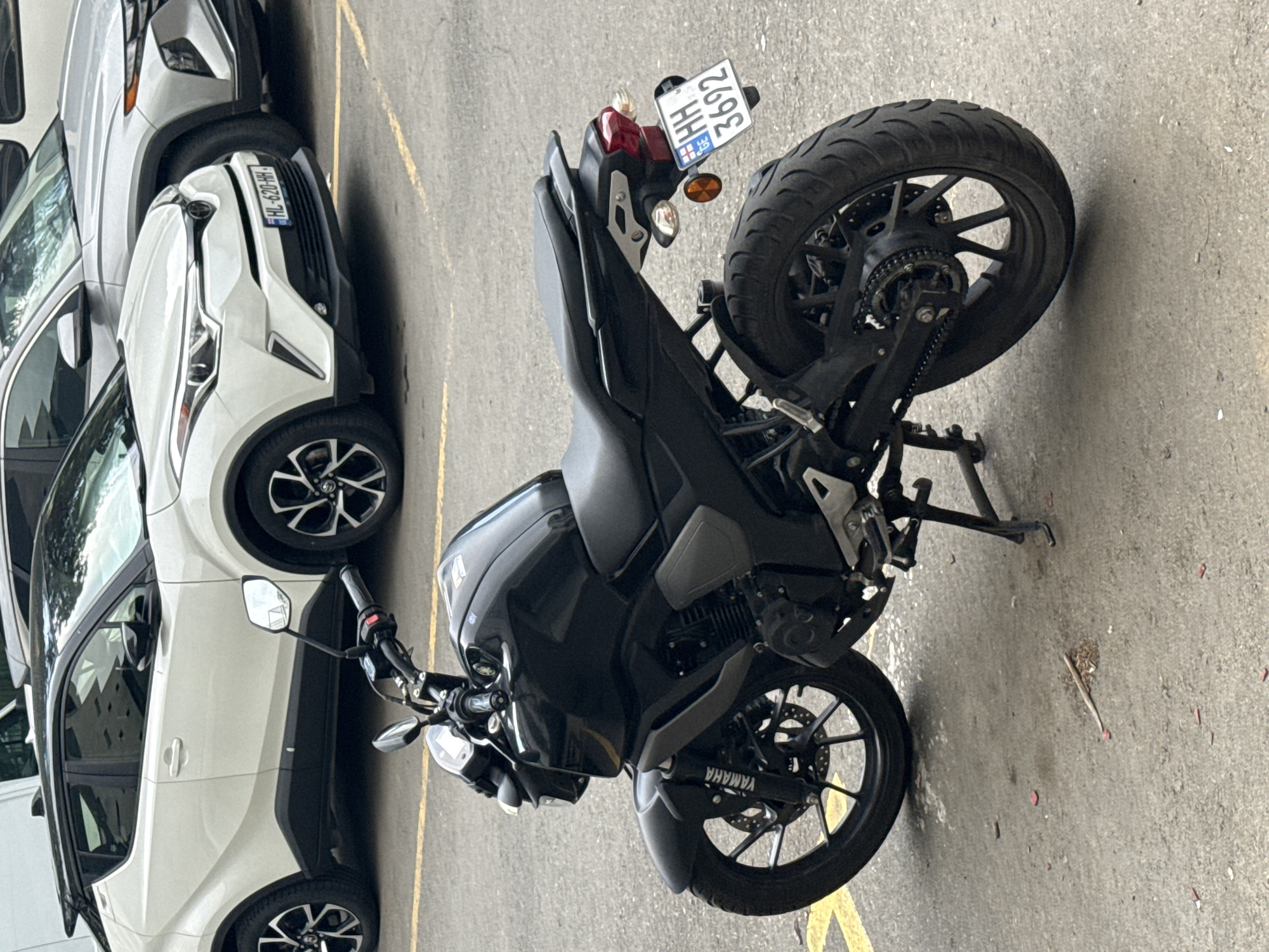 This is a media file of a parked black Yamaha motorcycle in Tbilisi, Georgia. The motorcycle is the primary subject and is positioned in the foreground, angled slightly to the right. Its details, including the engine, wheels, and handlebars, are clearly visible. The license plate of the motorcycle reads "HH 2692".
In the background, several cars are parked. To the left of the motorcycle, a white Toyota SUV is visible, with its front bumper and headlights in view. Part of its license plate, which reads "WL-029-HH", can also be seen. Further to the left, another car is partially visible. The scene appears to be an outdoor parking lot, indicated by the asphalt ground and yellow painted lines.
The image is taken from a high-angle perspective, looking down on the motorcycle and cars. The lighting suggests it is daytime, and there are no indications of specific weather conditions. The overall scene is static, with no activity taking place beyond the parked vehicles. There is no visible text on the motorcycle other than the license plate and the "YAMAHA" branding on the swingarm.