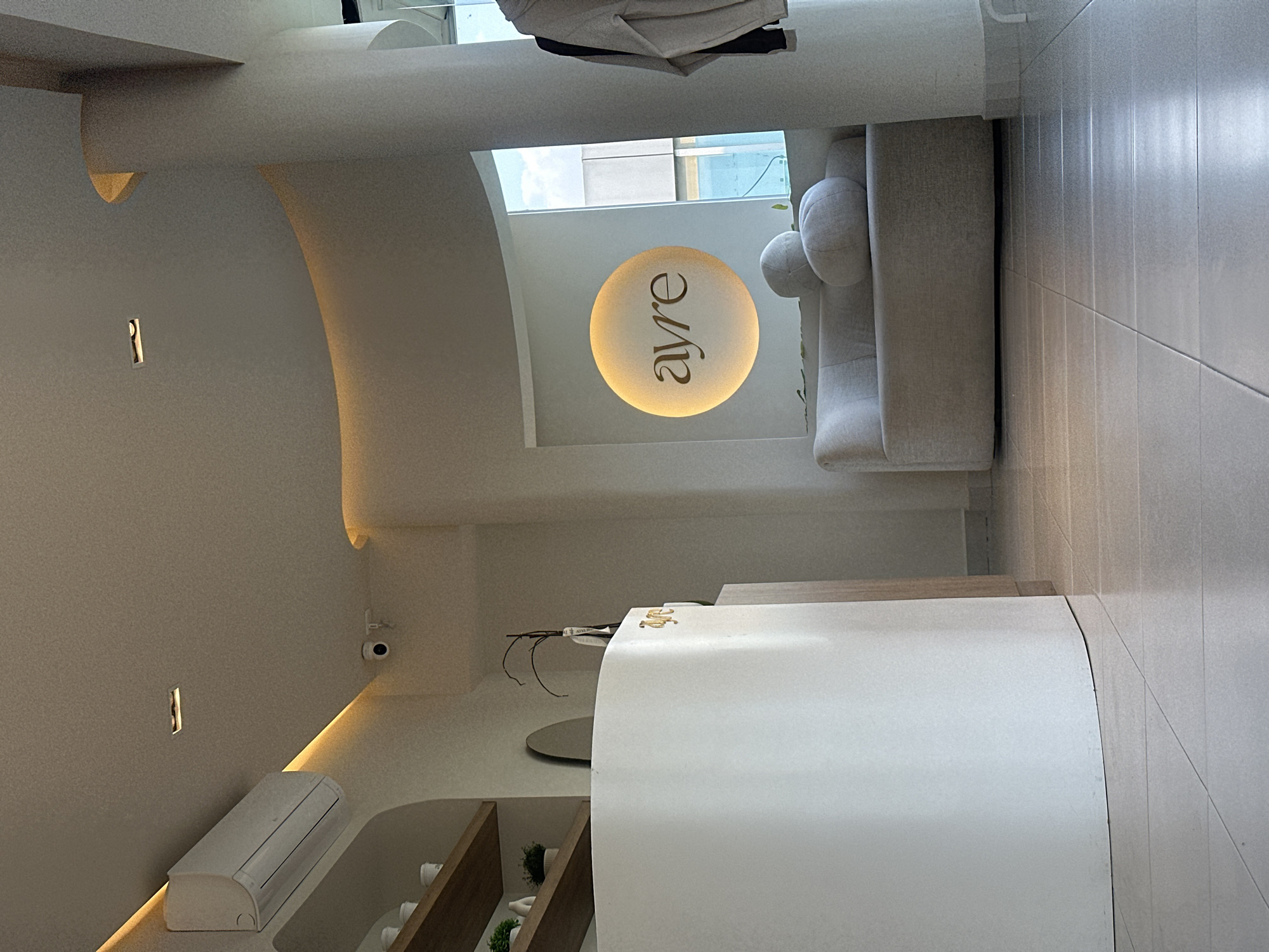 The image depicts an interior space. Dominating the right foreground is a white, curved structure, possibly a reception desk or counter, with its edge meeting a light-colored tiled floor. Behind the desk is a light-colored sofa with rounded cushions. Above the sofa, hanging from the ceiling is a circular sign with the word "ayre" on it.
On the left side of the image, the wall is painted a light color and features curved architectural elements, with some areas lit by recessed lighting. Visible on the wall are a small air conditioning unit and a security camera. Beneath the desk, shelves display decorative items, including small potted plants.
The location appears to be in an urban environment, as hinted by the view through the skylight above the sofa, showing parts of buildings and the sky. However, without additional visual information, pinpointing the exact location in San Bernardino Tlaxcalancingo, Mexico, is not possible. The scene is devoid of people.