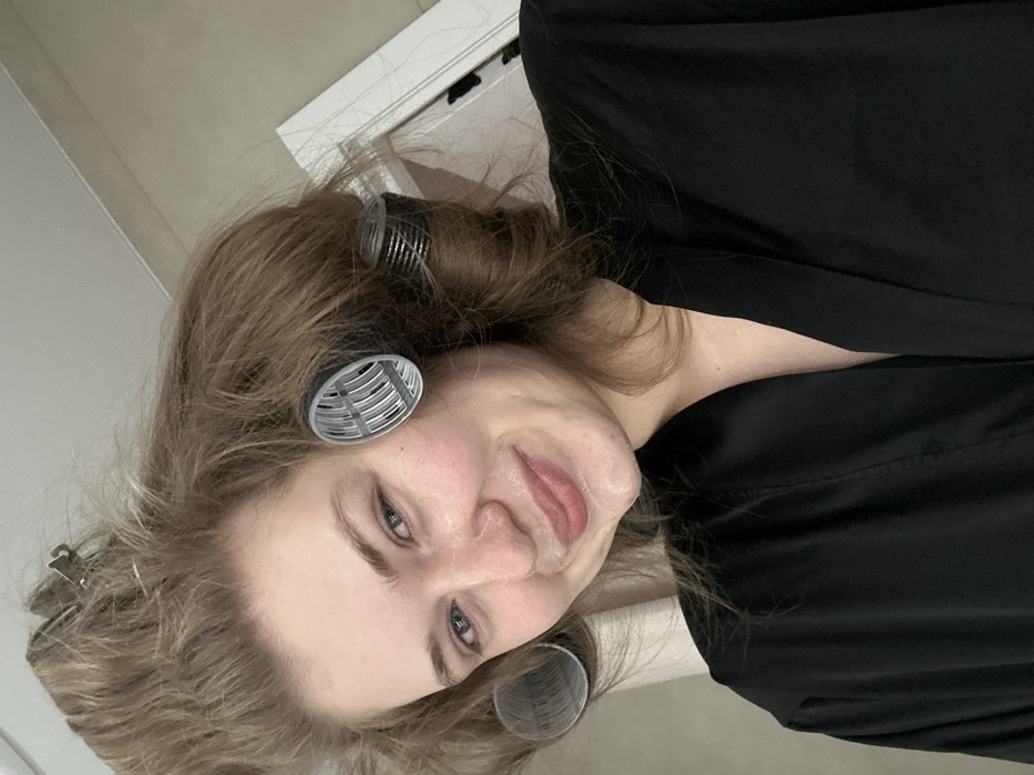 A woman with fair skin smiles at the camera. She has hair rollers in her blond hair. The woman is wearing a black robe, possibly satin. A white doorframe is in the background, and the rest of the image appears to be in a bathroom or dressing room setting. The image is rotated sideways.