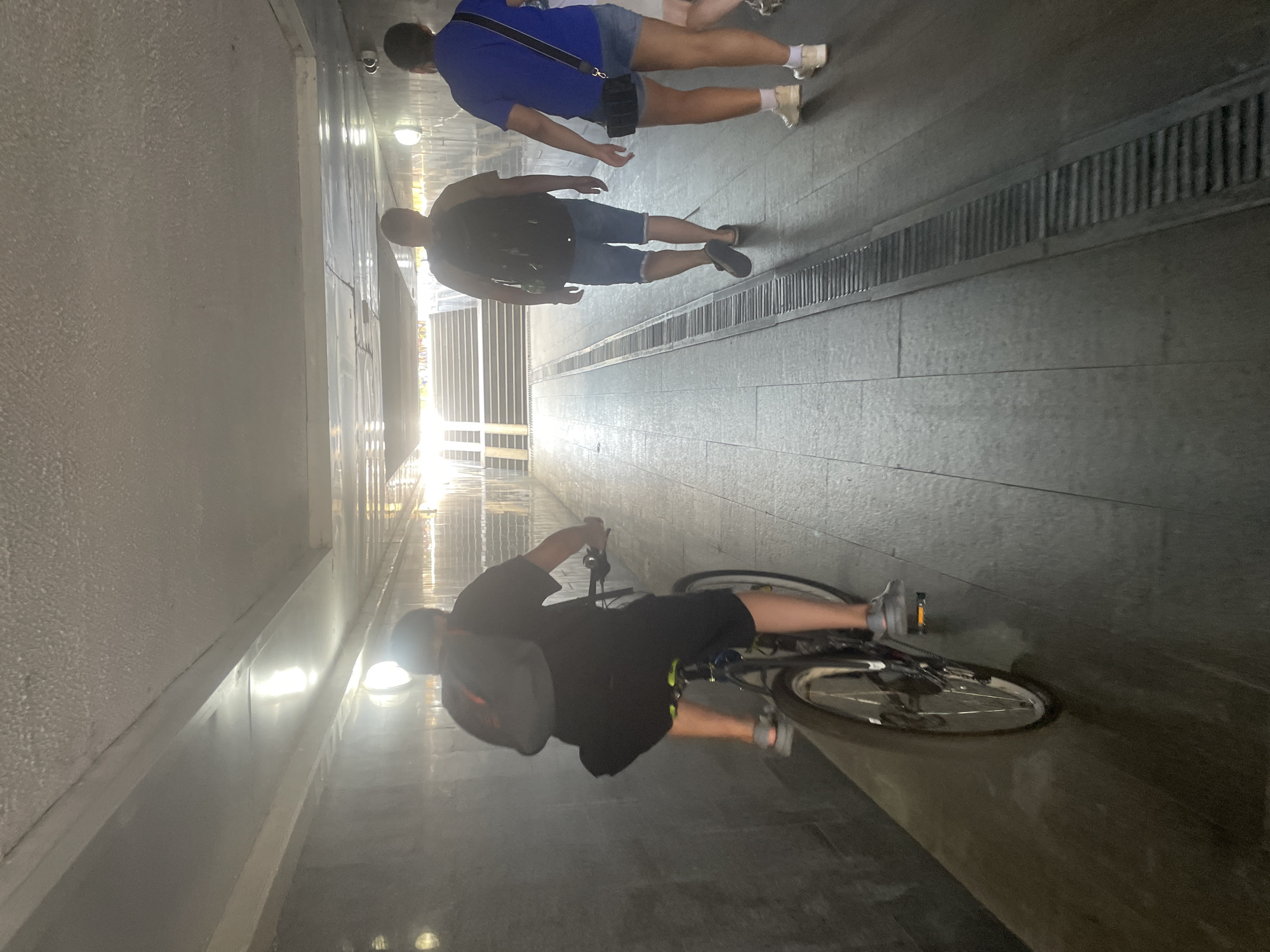 In a brightly lit pedestrian underpass in Sochi, Russia, a person on a bicycle rides away from the camera. The cyclist, wearing a black t-shirt, black shorts, and a grey backpack, is positioned in the left foreground of the image. The underpass has a tiled floor and grey, stone-like walls. Ahead, two individuals are walking away from the camera towards a set of stairs, one in a blue t-shirt and denim shorts, carrying a black bag, and the other in a tan t-shirt and denim shorts with a black backpack. The lighting suggests it is daytime, with sunlight streaming in from an opening at the end of the underpass where the stairs are located.