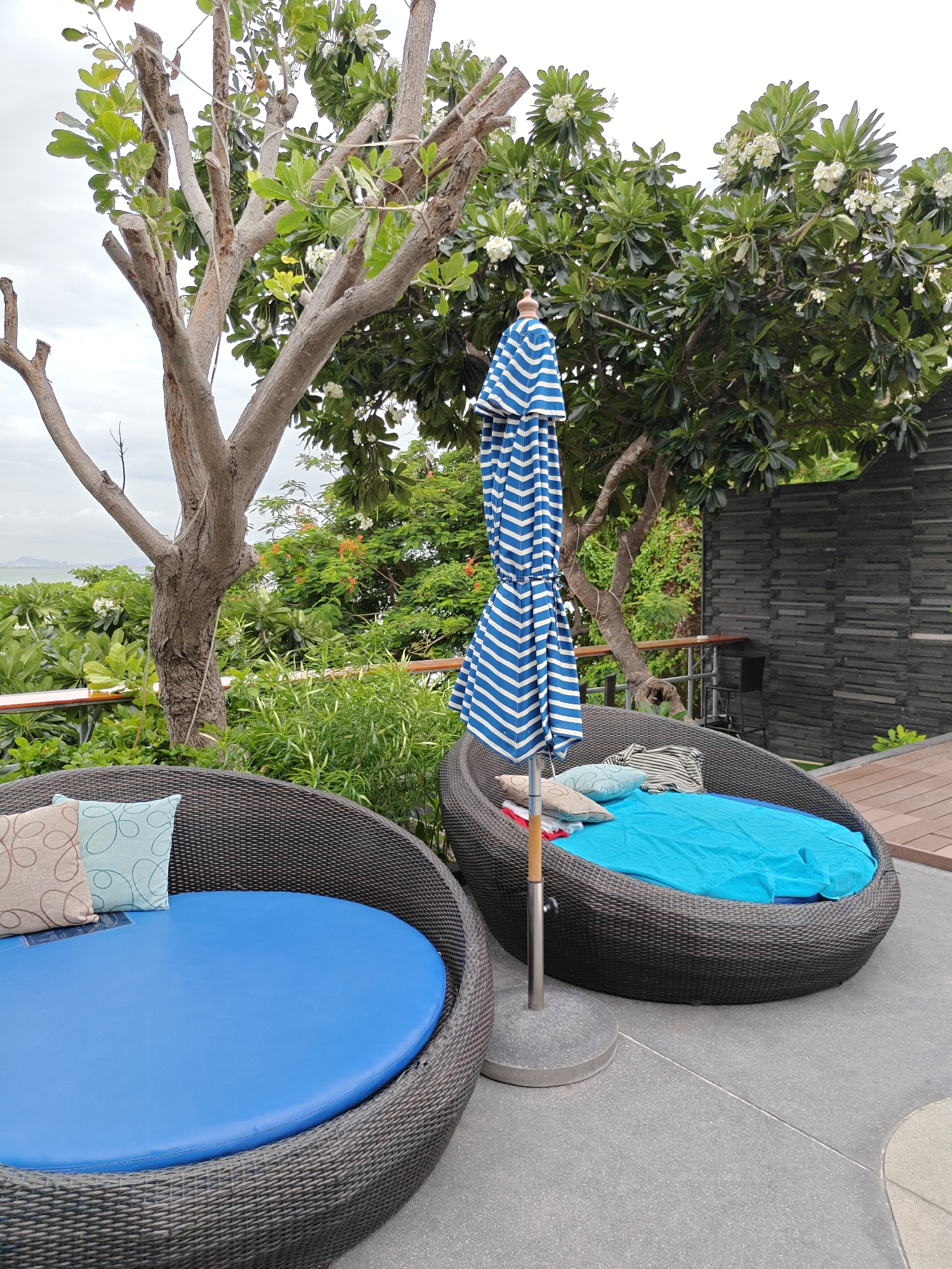 A wide shot captures an outdoor seating area with two large, round, woven furniture pieces with blue cushions on a grey paved area. A blue and white striped umbrella is partially open and situated between the two seating areas. In the background, lush green trees and foliage frame the scene. A stone wall with horizontal lines and a wooden deck are visible to the right. The ocean and distant landforms can be seen in the far background under a cloudy sky. The location is the city of Pattaya, Thailand. There are no people present in the image.